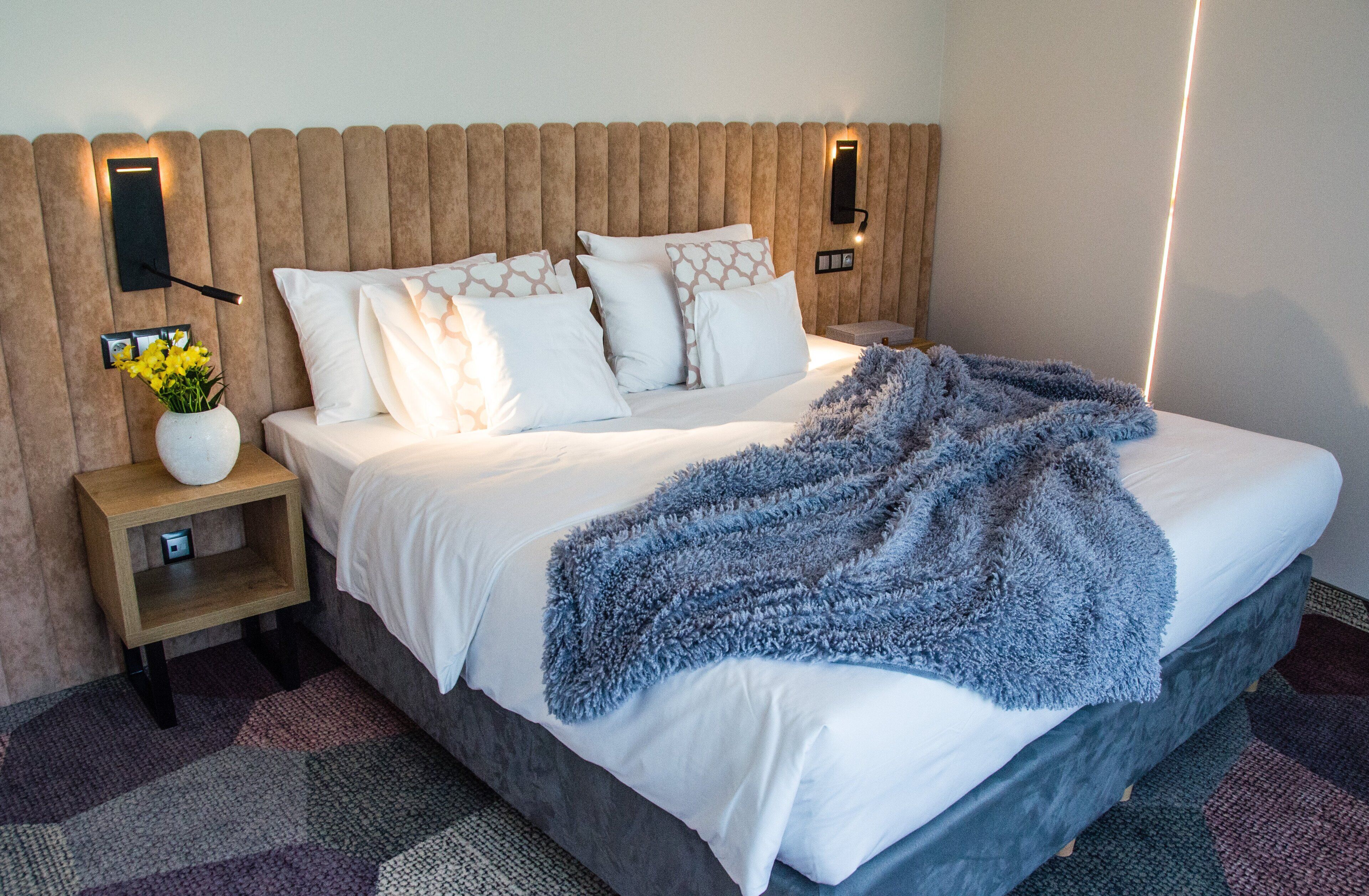 Una cama bien hecha con sábanas blancas y un edredón azul, una mesita de noche de madera con un florero y una lámpara de pared.