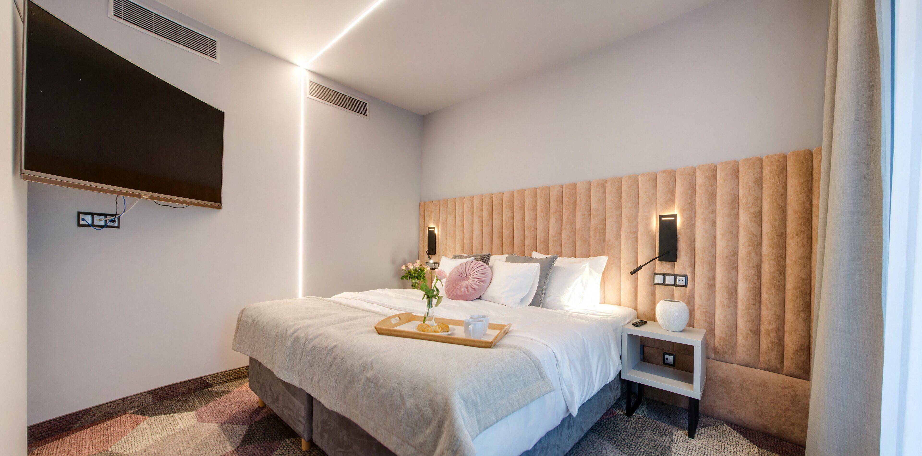 A hotel room with a bed, a bedside table, a TV mounted on the wall, and a wall-mounted air vent.