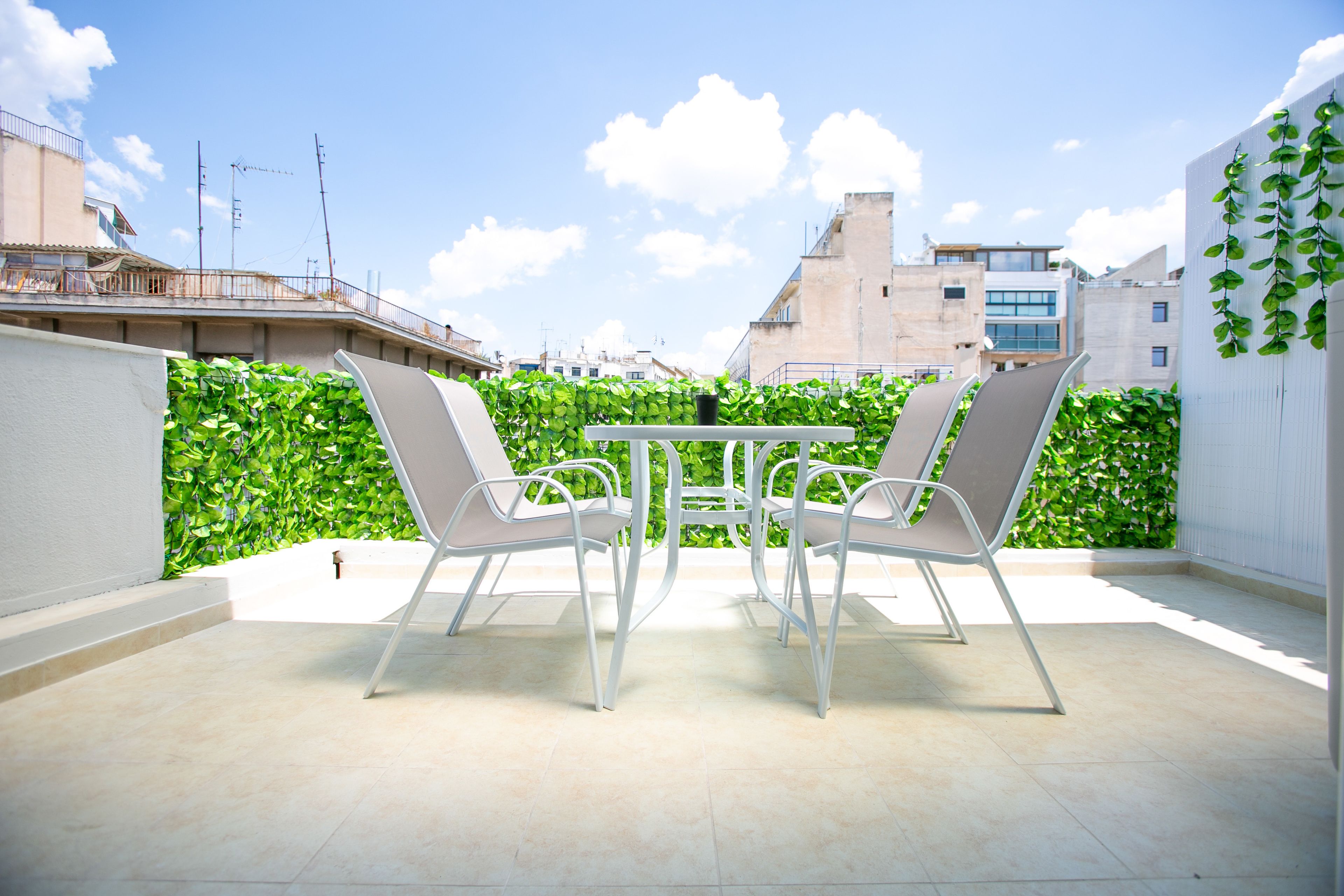 Eine Terrasse im Freien mit Tisch und vier Stühlen, umgeben von einer grünen Hecke und einer weißen Mauer.