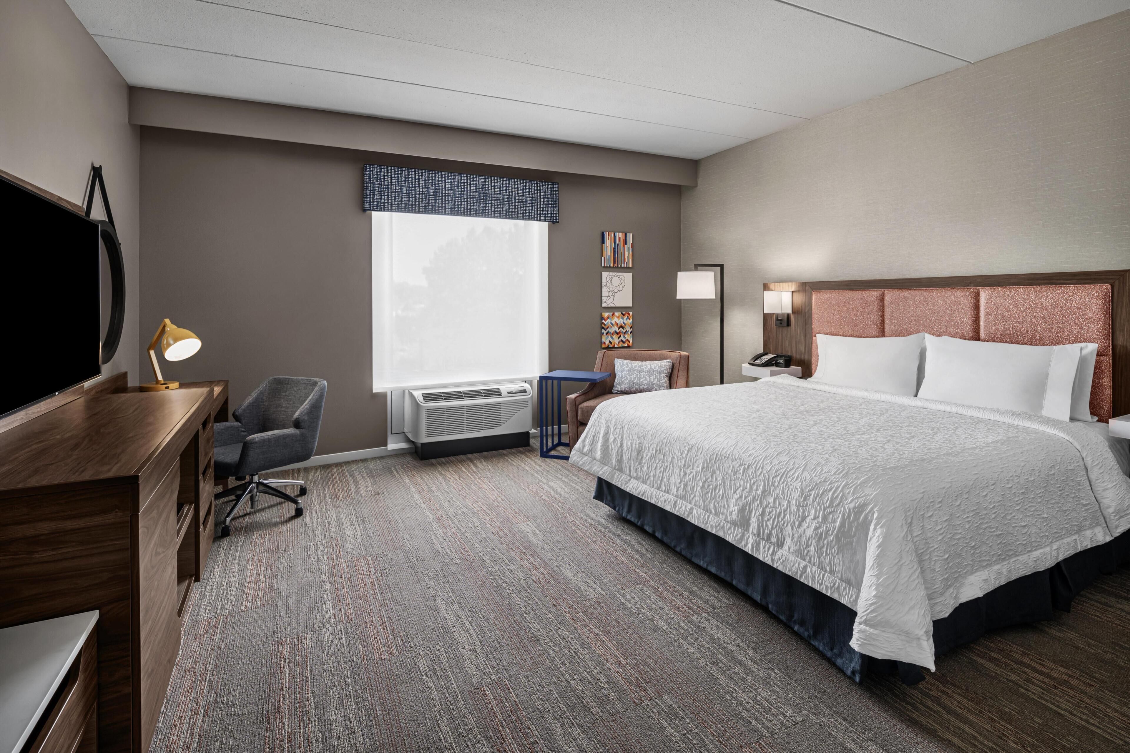 Habitación de hotel con una cama grande, un escritorio con silla, televisión, ventana con persianas y aire acondicionado instalado en la pared.