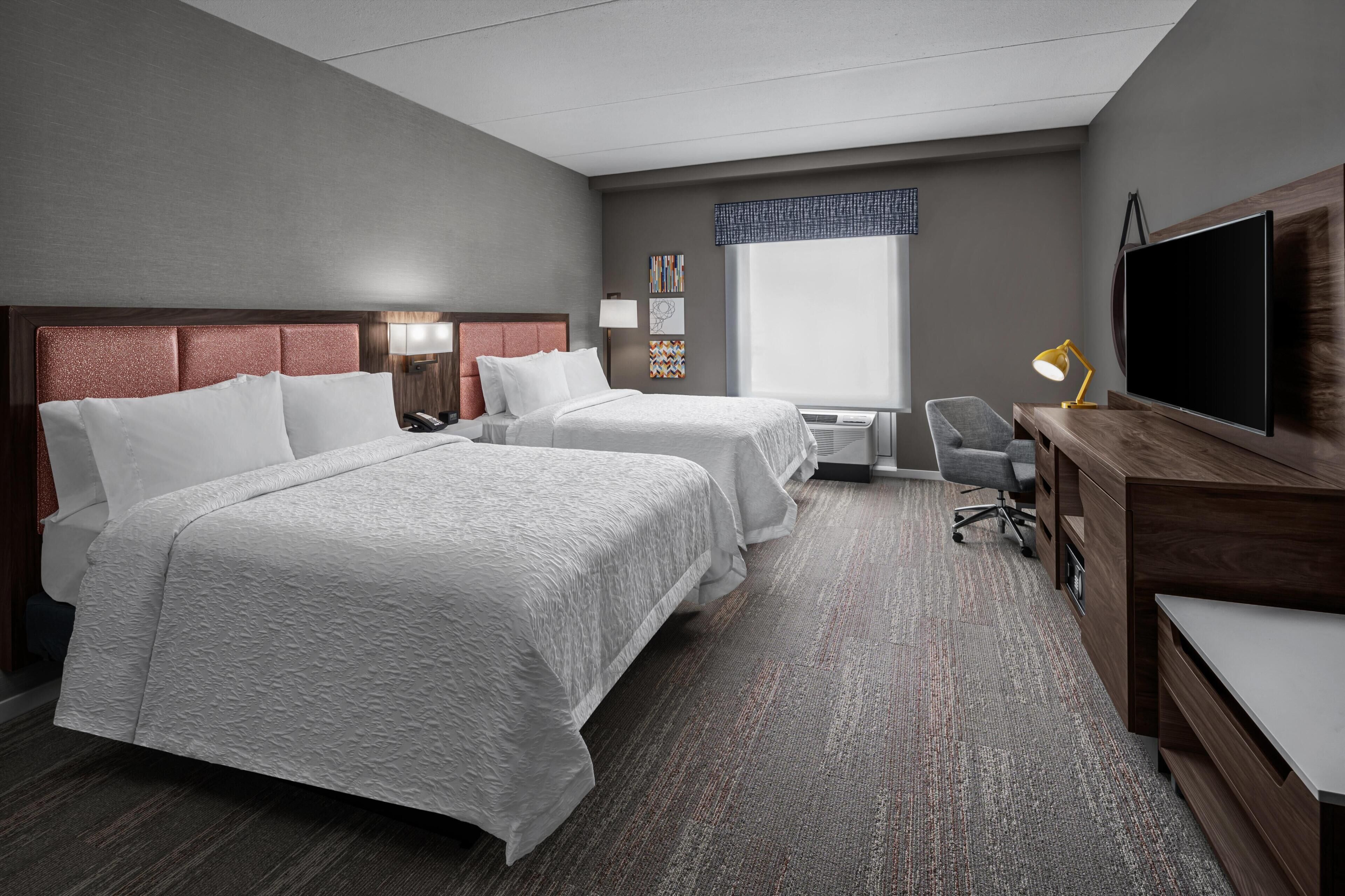 A hotel room with two beds, a desk, a TV, and a window.