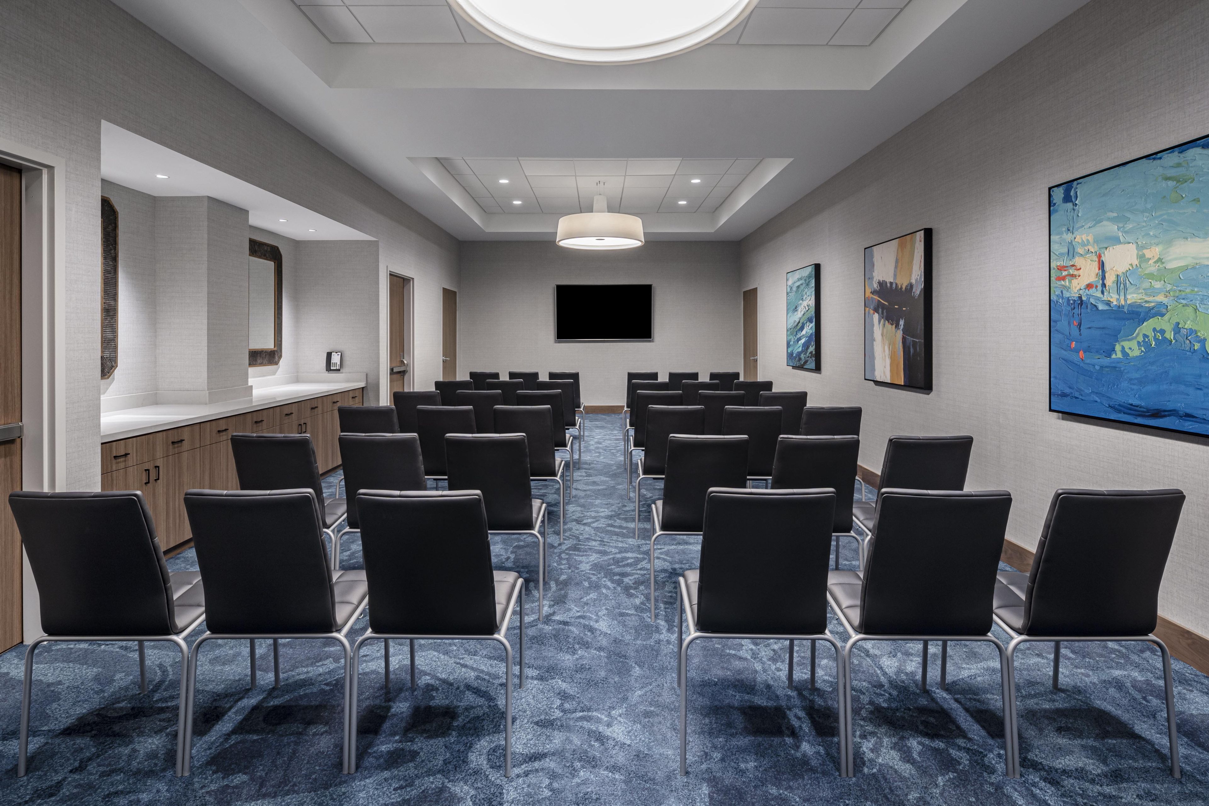 Un salón de conferencias con filas de sillas, una pantalla grande y obras de arte en las paredes.