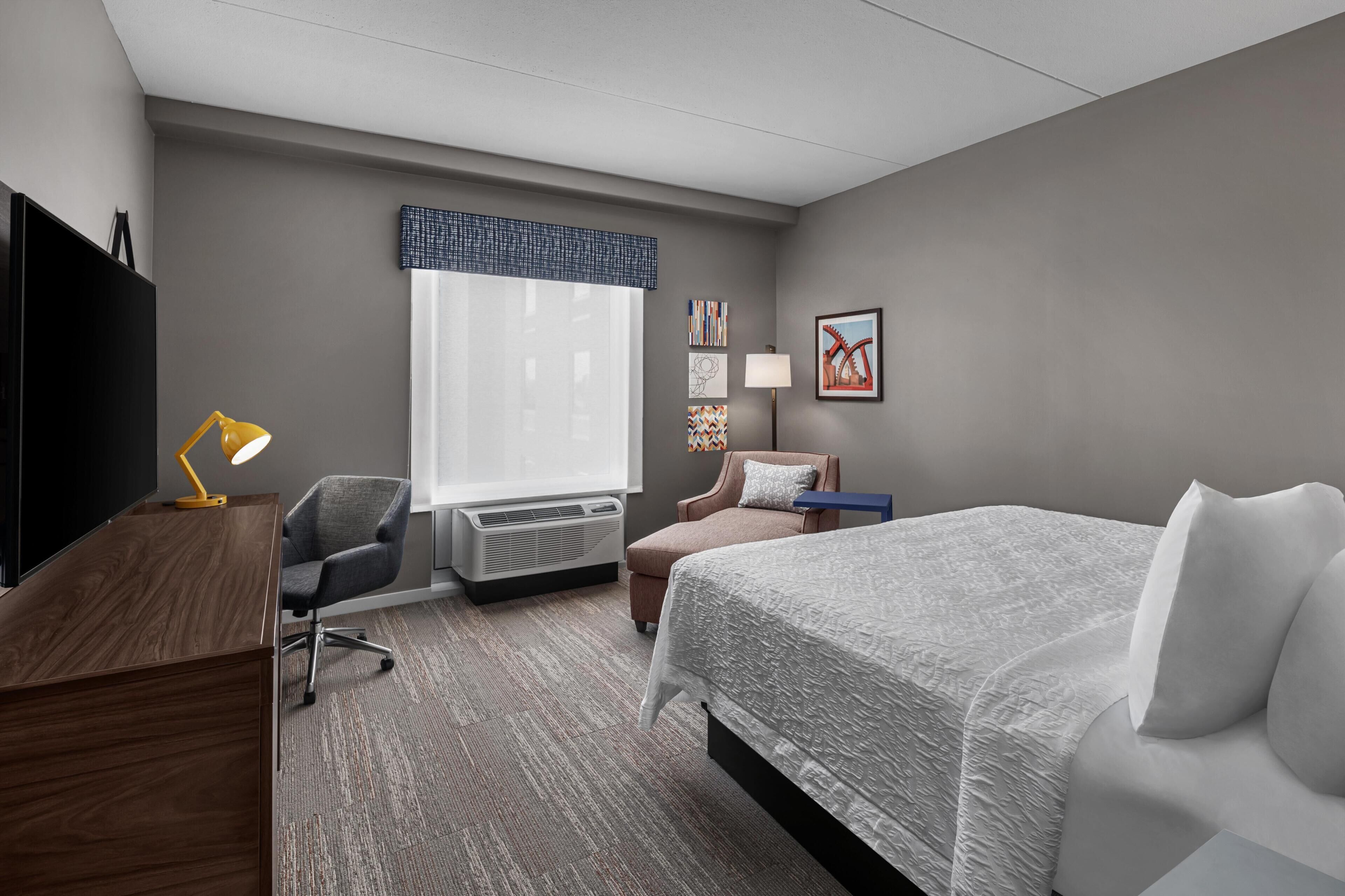 A hotel room with a bed, desk, chair, air conditioning unit, and a TV.