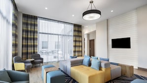 A modern hotel room with a sectional sofa, armchairs, a flat-screen TV, and large windows with curtains.