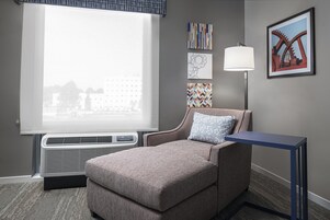 Habitación de hotel con un ventanal grande, un aire acondicionado instalado en la pared, un sofá gris con un cojín estampado, una mesita pequeña y cuadros en la pared.
