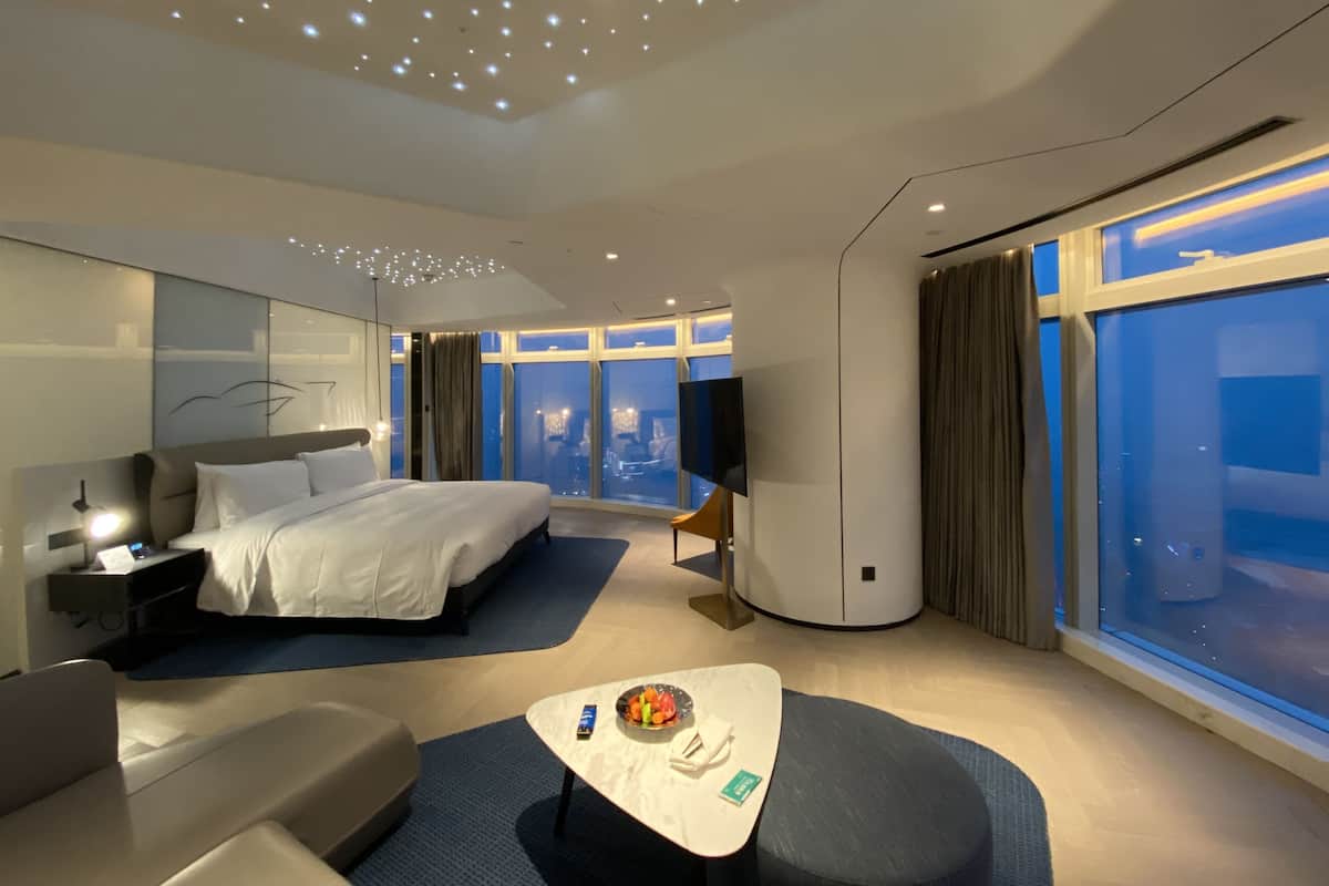 A modern hotel room with a large bed, a desk with a lamp, a small table with a bowl of fruit, and a large window offering a city view.