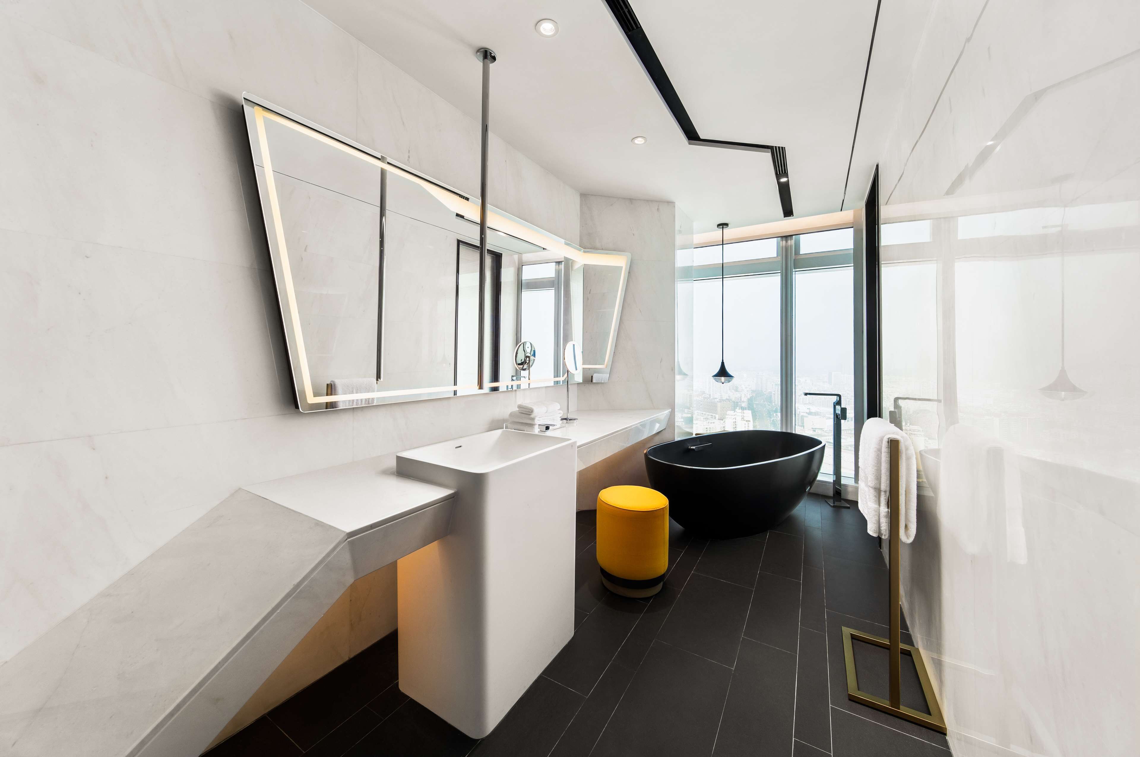 Un baño moderno con un espejo grande, un lavabo blanco, una bañera negra y un taburete amarillo.