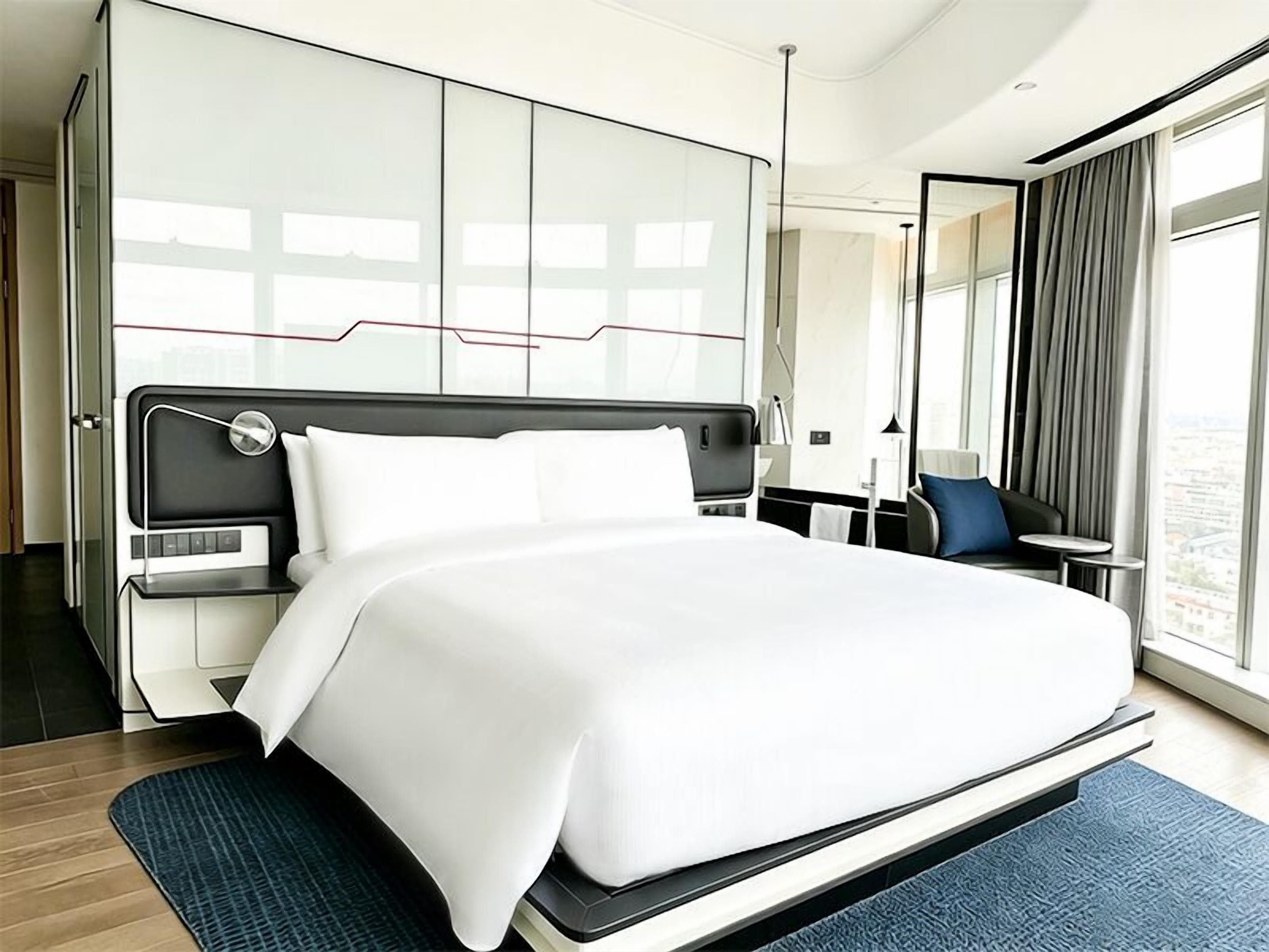 A modern hotel room with a large bed, a desk, a chair, and a view of the city.