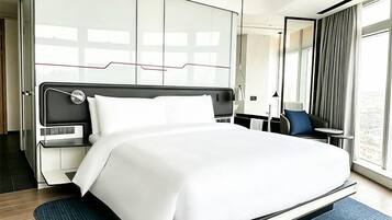 A modern hotel room with a large bed, a desk, a chair, and a view of the city.
