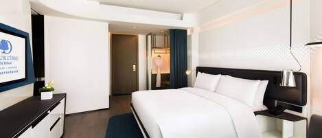 A modern hotel room with a neatly made bed, a desk with a lamp, and a view of the hallway.