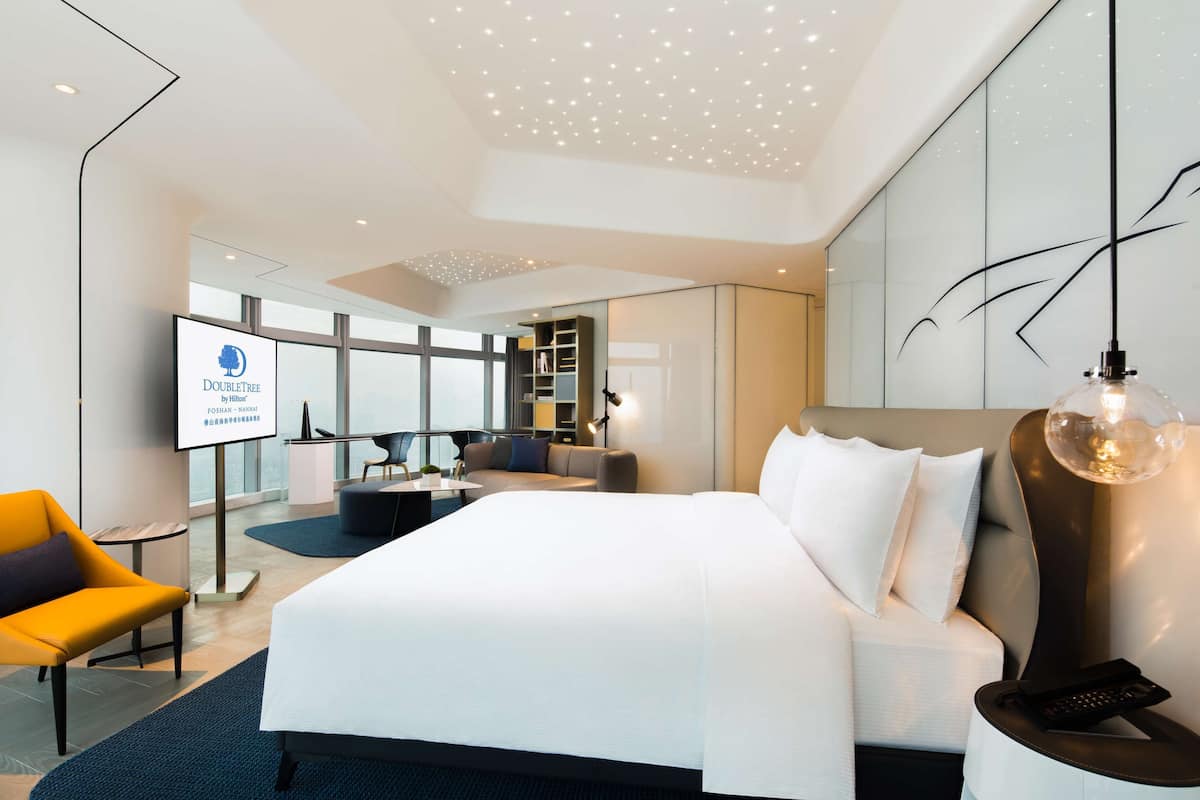 A modern hotel room with a large bed, a desk, a chair, and a TV displaying the DoubleTree logo.