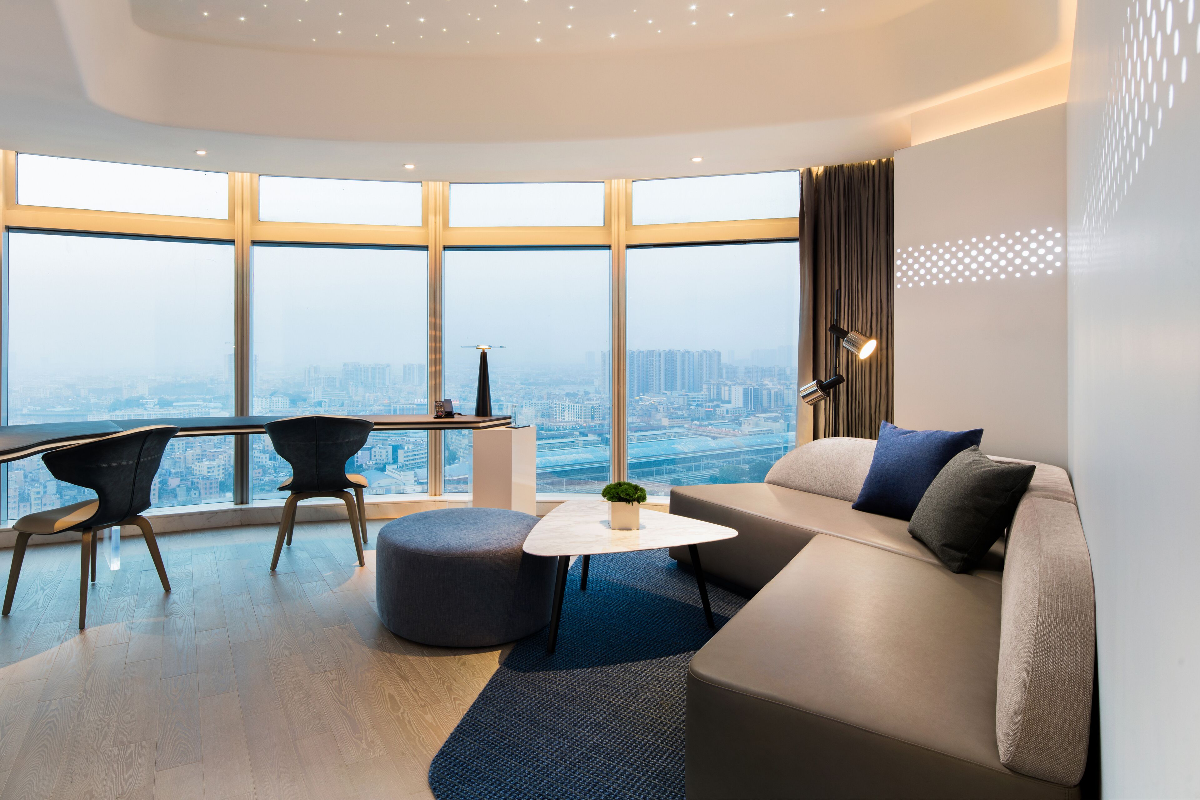 A modern living room with a large window, a sofa, a coffee table, and a desk with chairs.