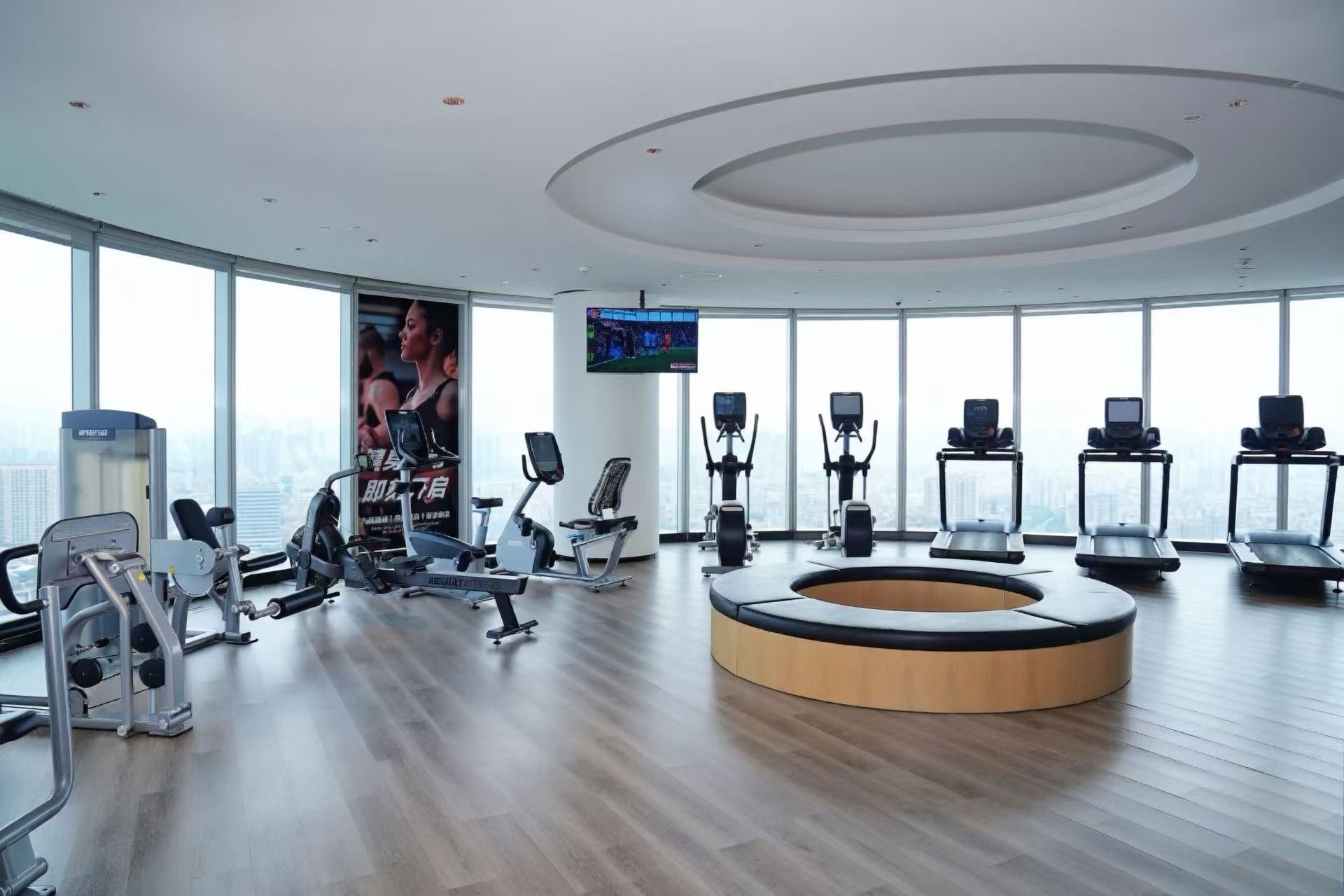 A modern gym with cardio equipment, weight machines, and a large window offering a city view.