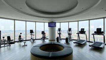 A modern gym with a panoramic city view, equipped with various cardio machines and a central circular seating area.