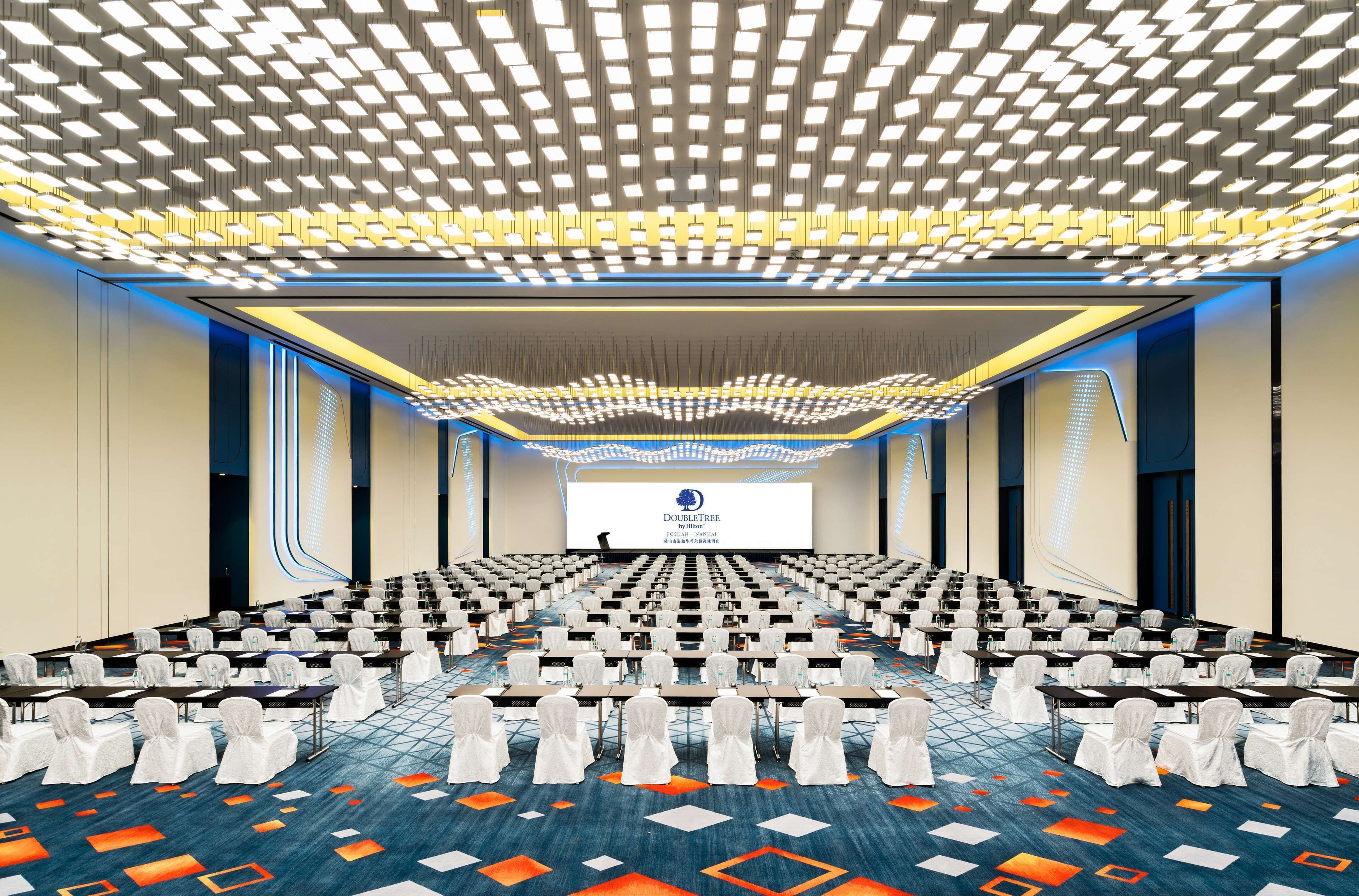 A conference room with rows of tables and chairs, a large screen at the front, and a patterned ceiling.