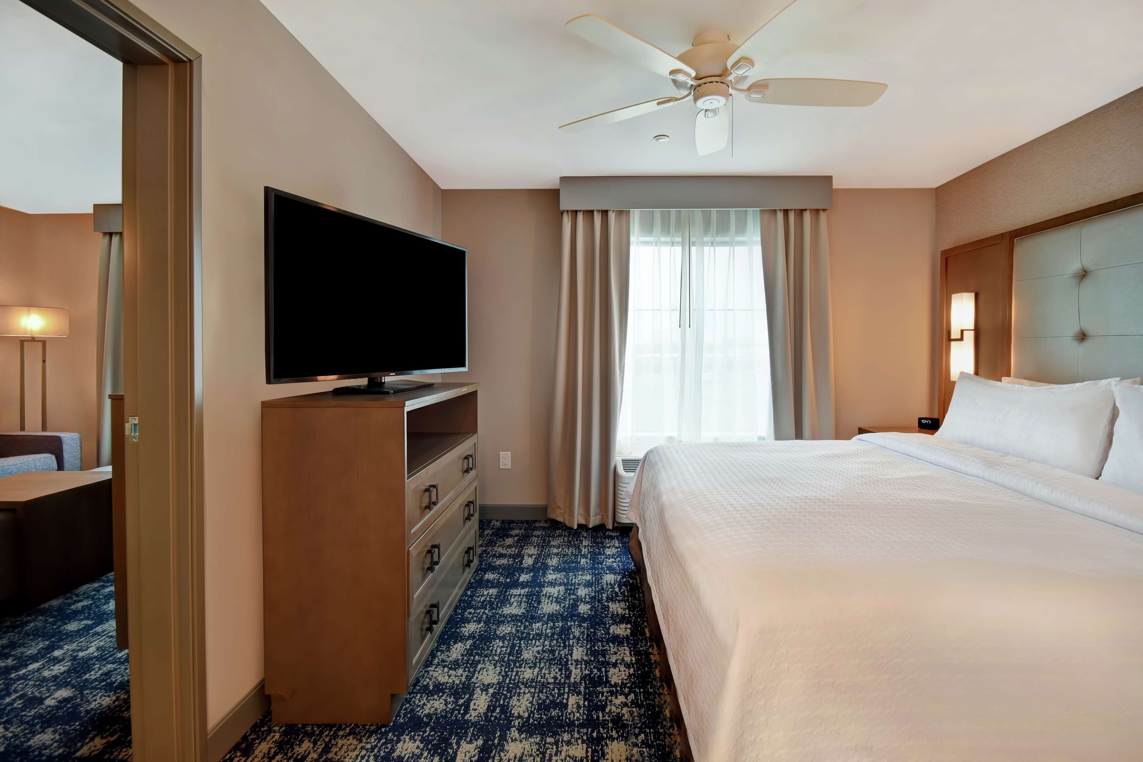 A hotel room with a bed, a television on a wooden dresser, a ceiling fan, and a window with curtains.