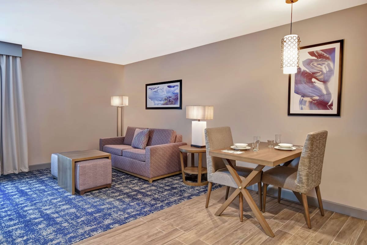 A modern hotel room with a sofa, a small table, a dining table with chairs, and two floor lamps.