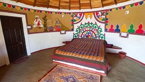 A room with a bed, a rug, and a wall with traditional artwork. - Banni Village Stay (Bhuj)