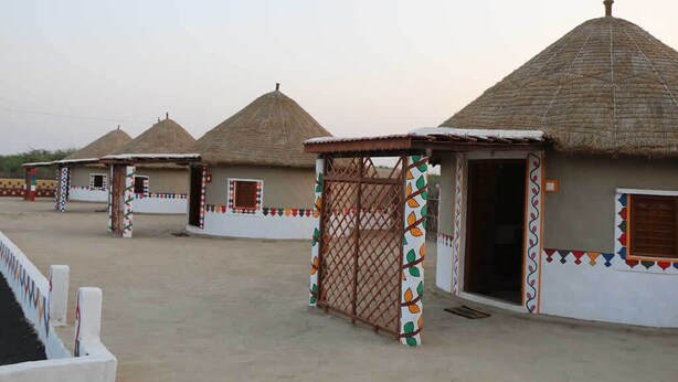 A row of traditional thatched-roof huts with decorative patterns and a gated entrance. - Banni Village Stay (Bhuj)