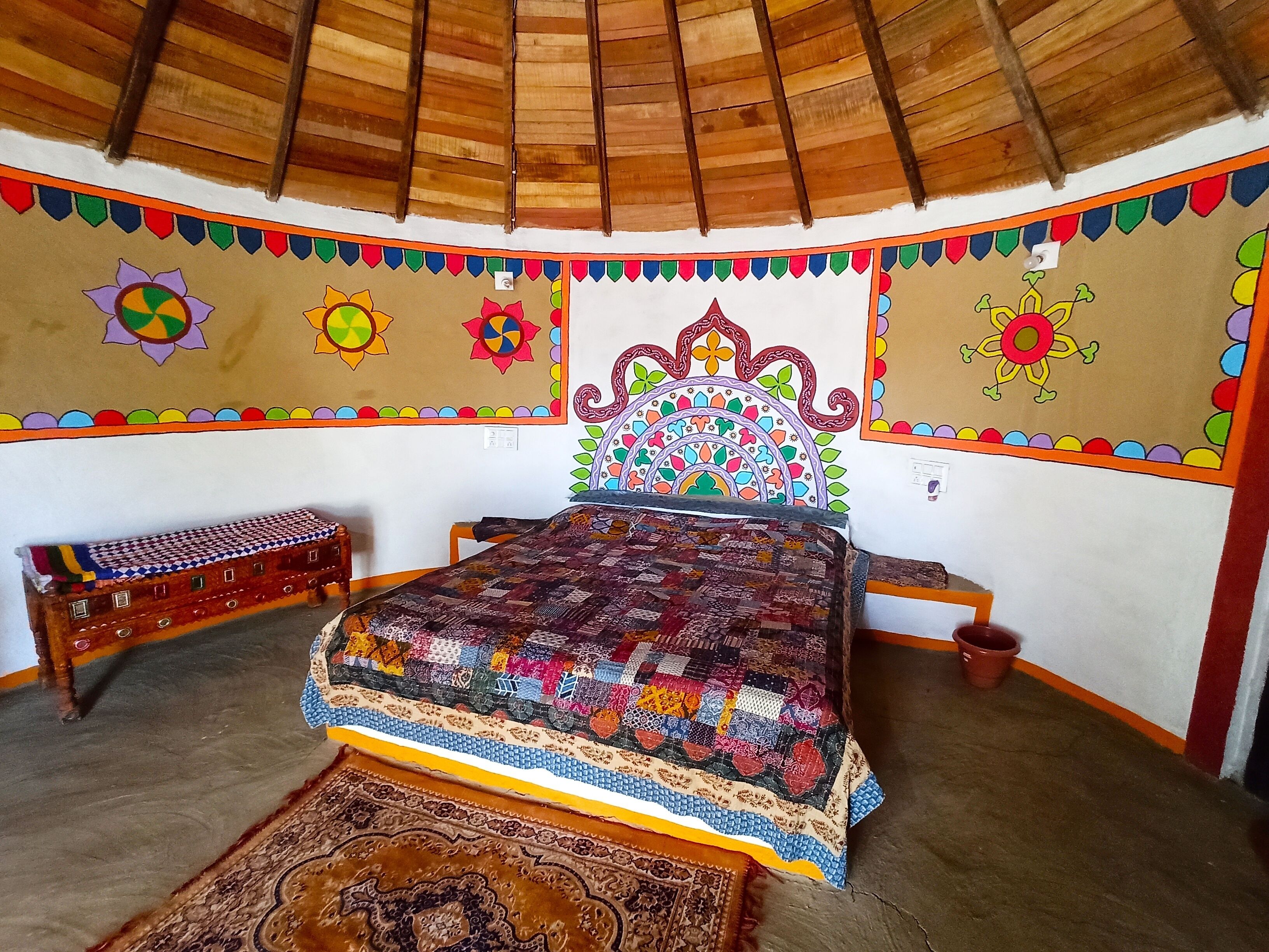 Una habitación con techo de madera, una cama con colcha de colores, un tapete estampado y una pared decorada con motivos florales coloridos.