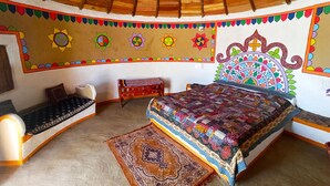 A room with a wooden ceiling, a bed with a colorful quilt, a patterned rug, and a small table with a cushion. - Banni Village Stay (Bhuj)