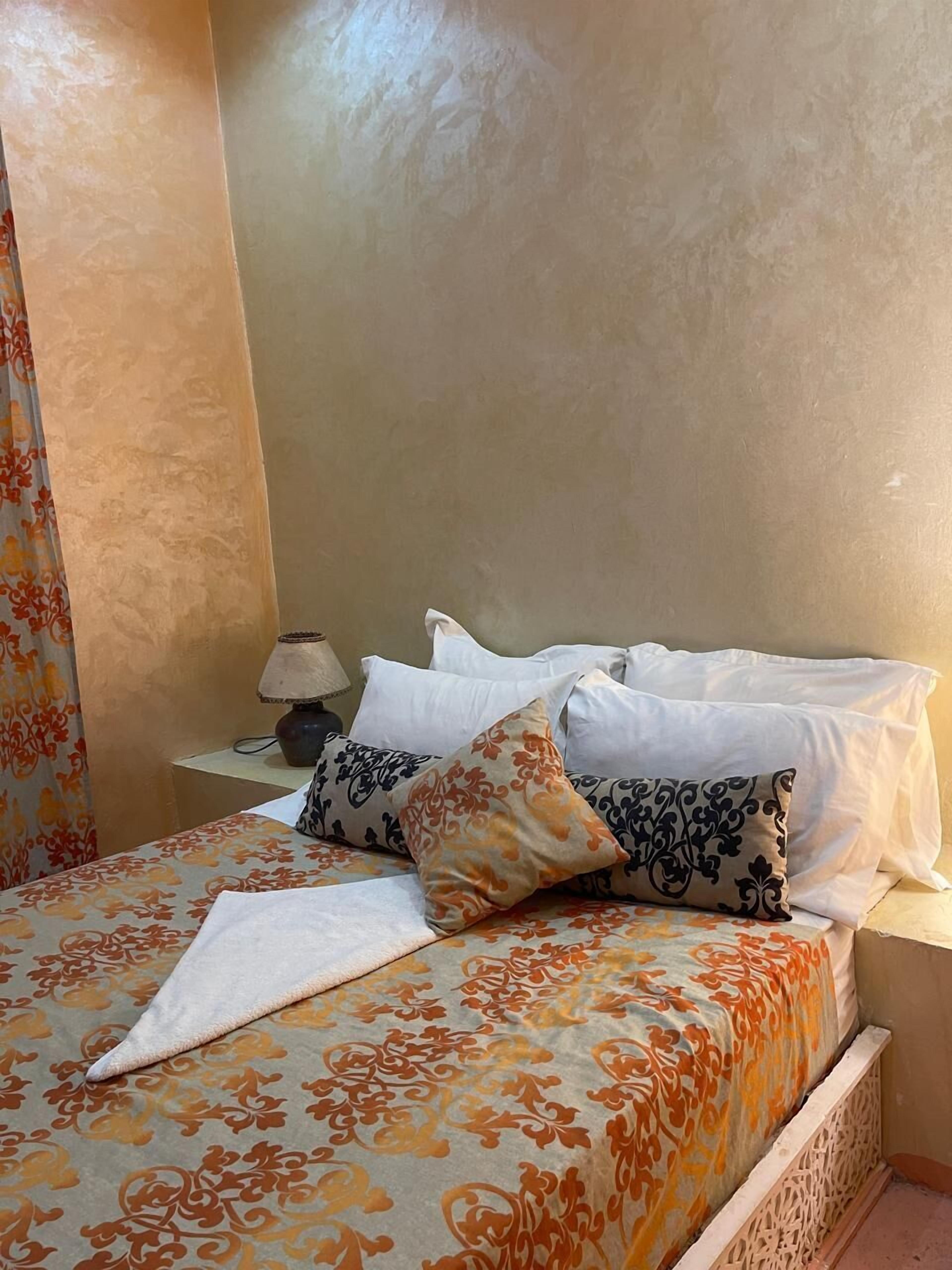 A neatly made bed with patterned bedding and pillows, a bedside table with a lamp, and a curtain with a floral design.