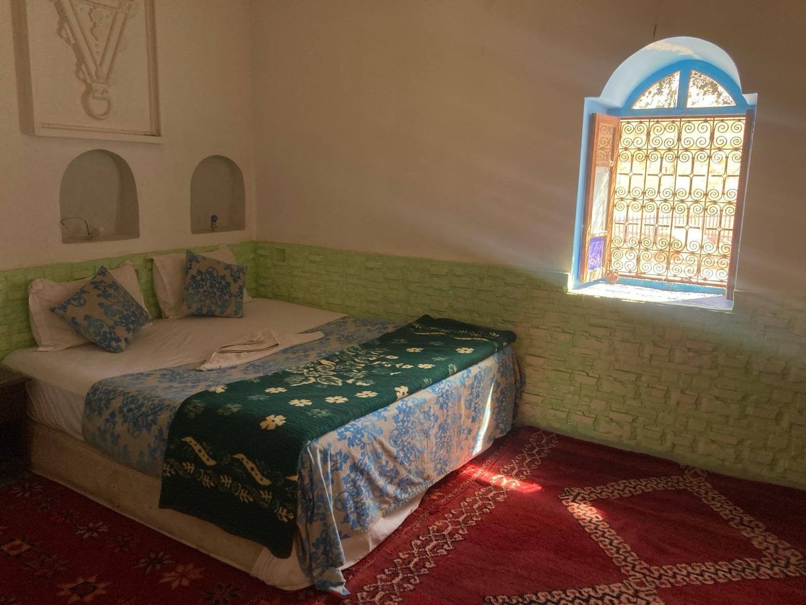 Una habitación con cama, ventana con celosías decorativas y alfombra estampada.