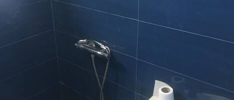 Une salle de bain avec des carreaux bleu foncé, une douche équipée d’un pommeau de douche à main, des toilettes et un sol imitant la pierre.