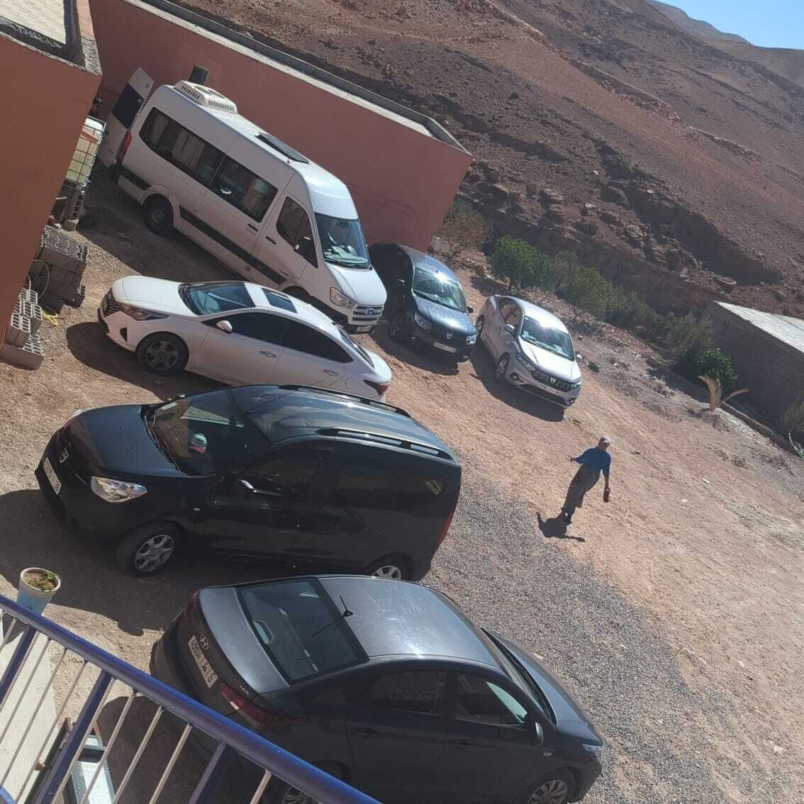 Un estacionamiento con varios coches, una furgoneta, una persona caminando y un edificio con tejado rojo.