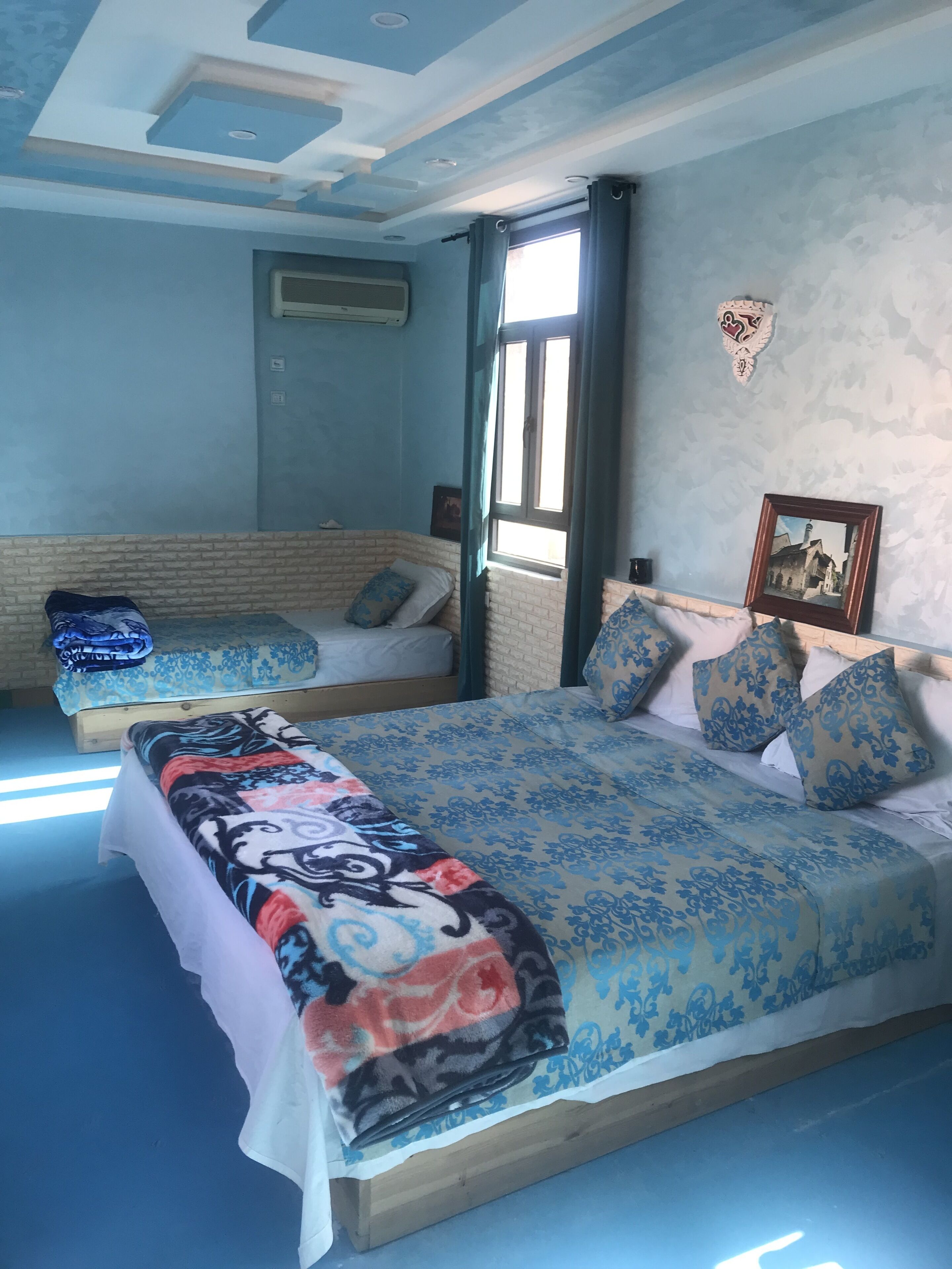 A bedroom with two beds, a brick wall, a window with curtains, and a picture on the wall.