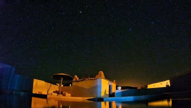 Vista noturna de uma propriedade à beira-mar, com estruturas iluminadas e reflexo nítido na água.
