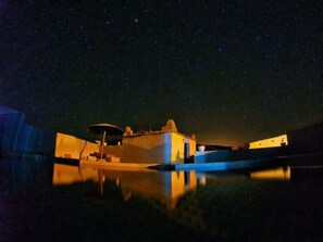Vista nocturna de una propiedad frente al agua, con edificios iluminados y un reflejo claro en el agua.