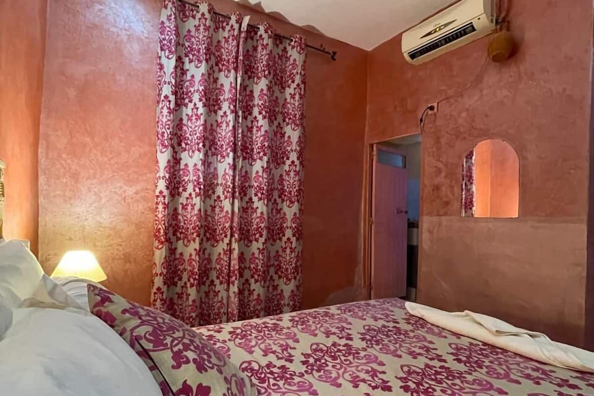 A bedroom with a bed, a nightstand, a lamp, a window with curtains, and an air conditioning unit.