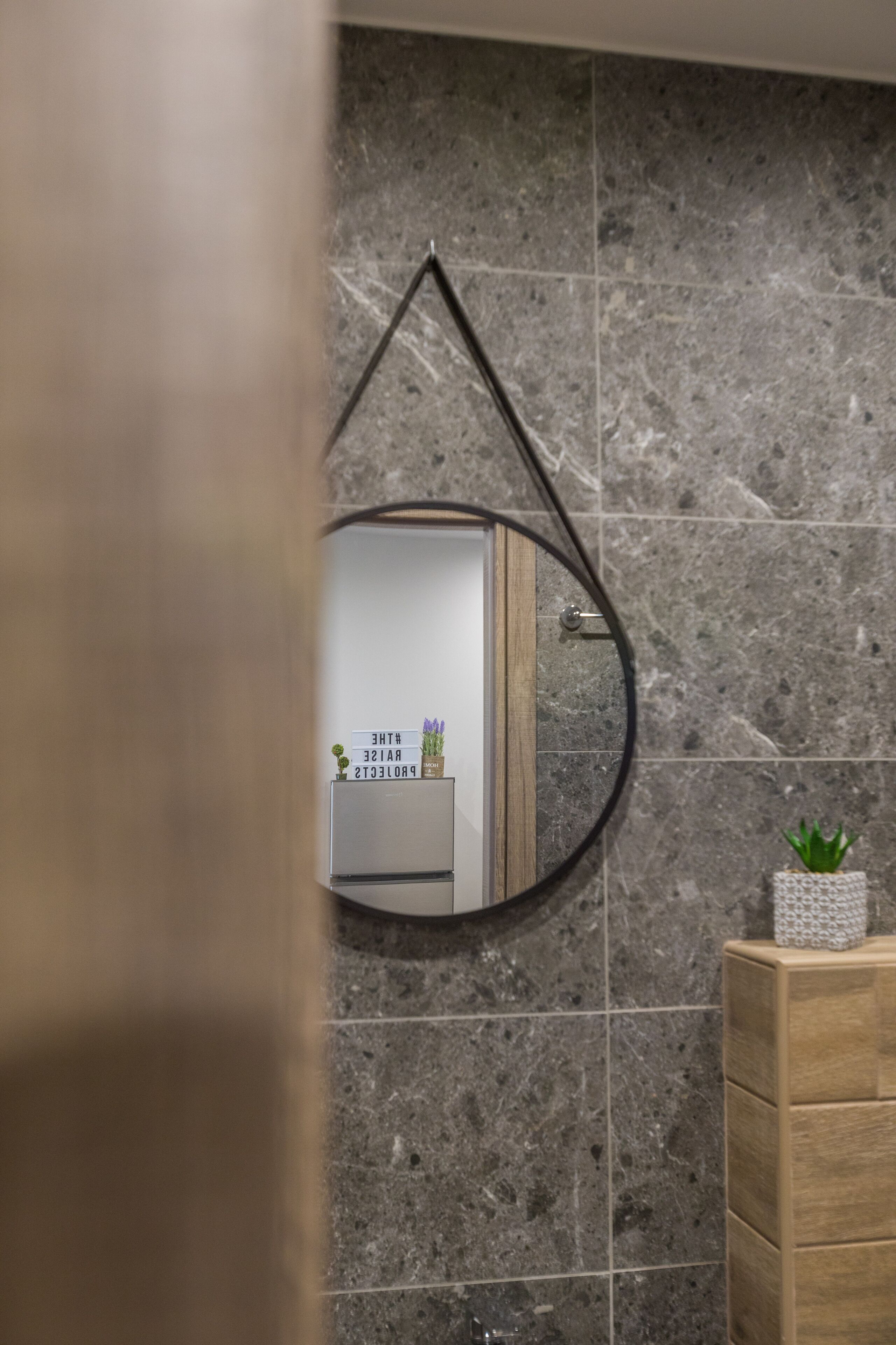 A bathroom with a marble wall, a wooden vanity, and a mirror with a hanging frame.