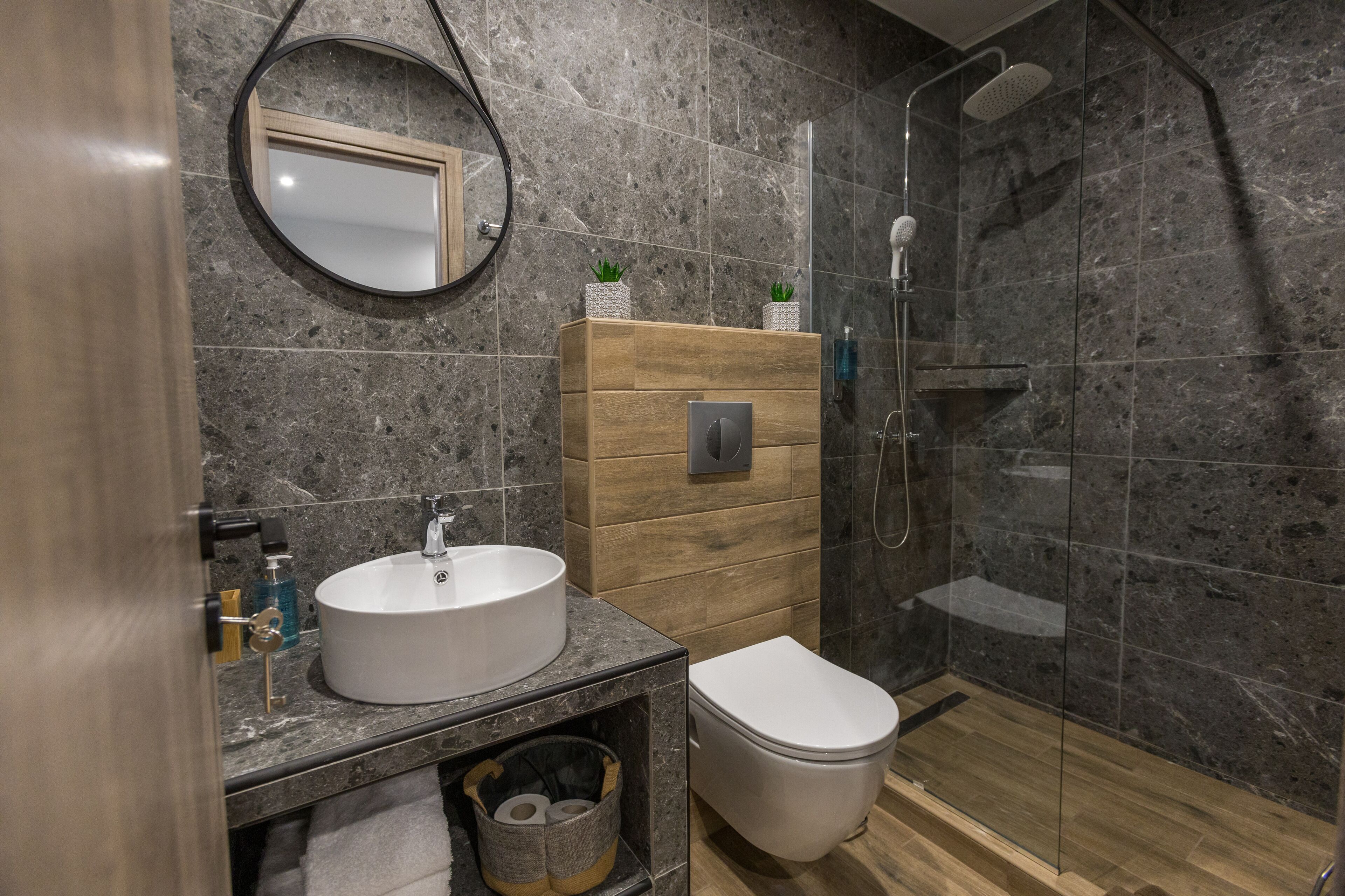A modern bathroom with a round mirror, a wooden vanity, a white sink, a toilet, and a glass-enclosed shower area.