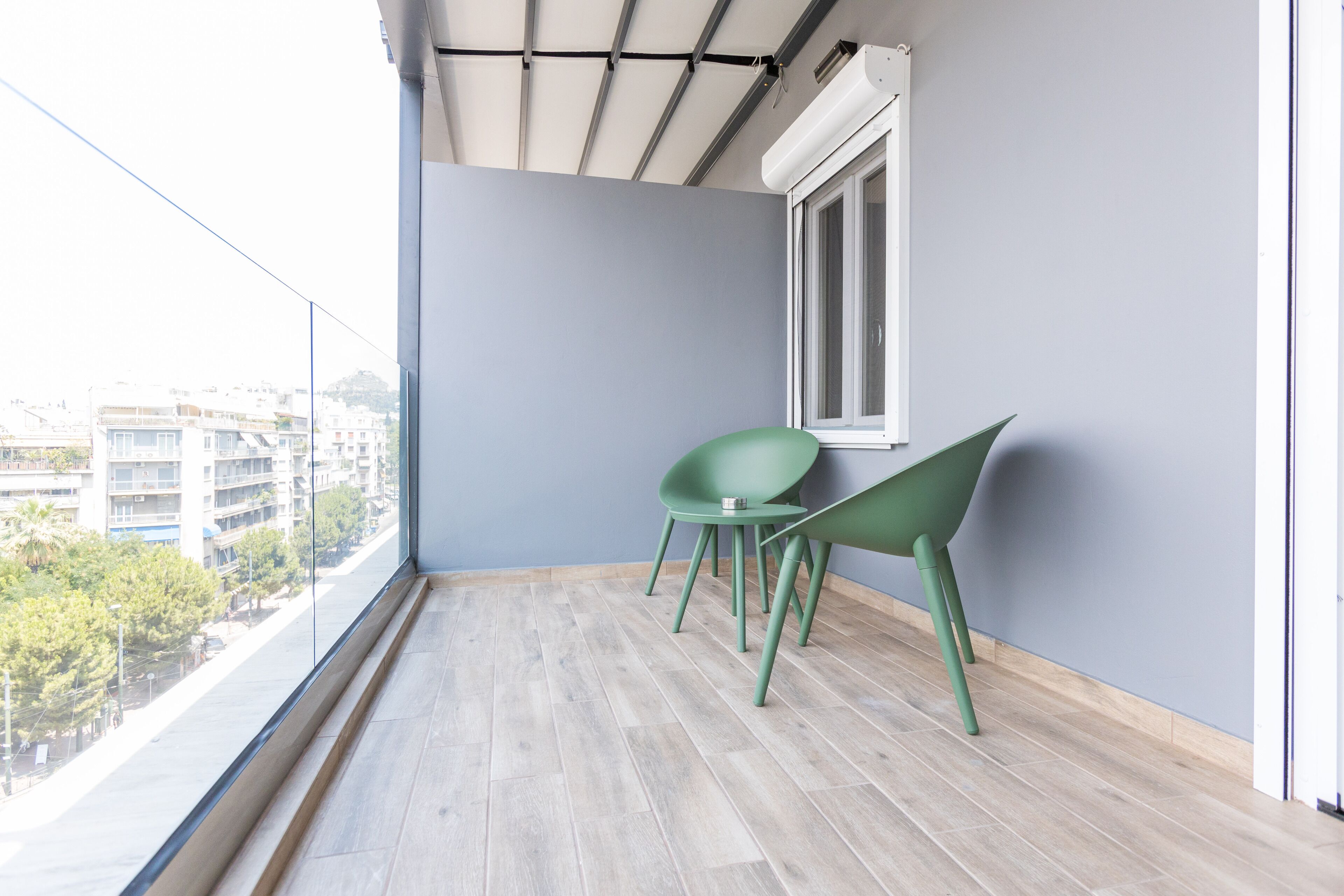 Un balcon donnant sur des immeubles et des arbres, une fenêtre et deux chaises vertes.