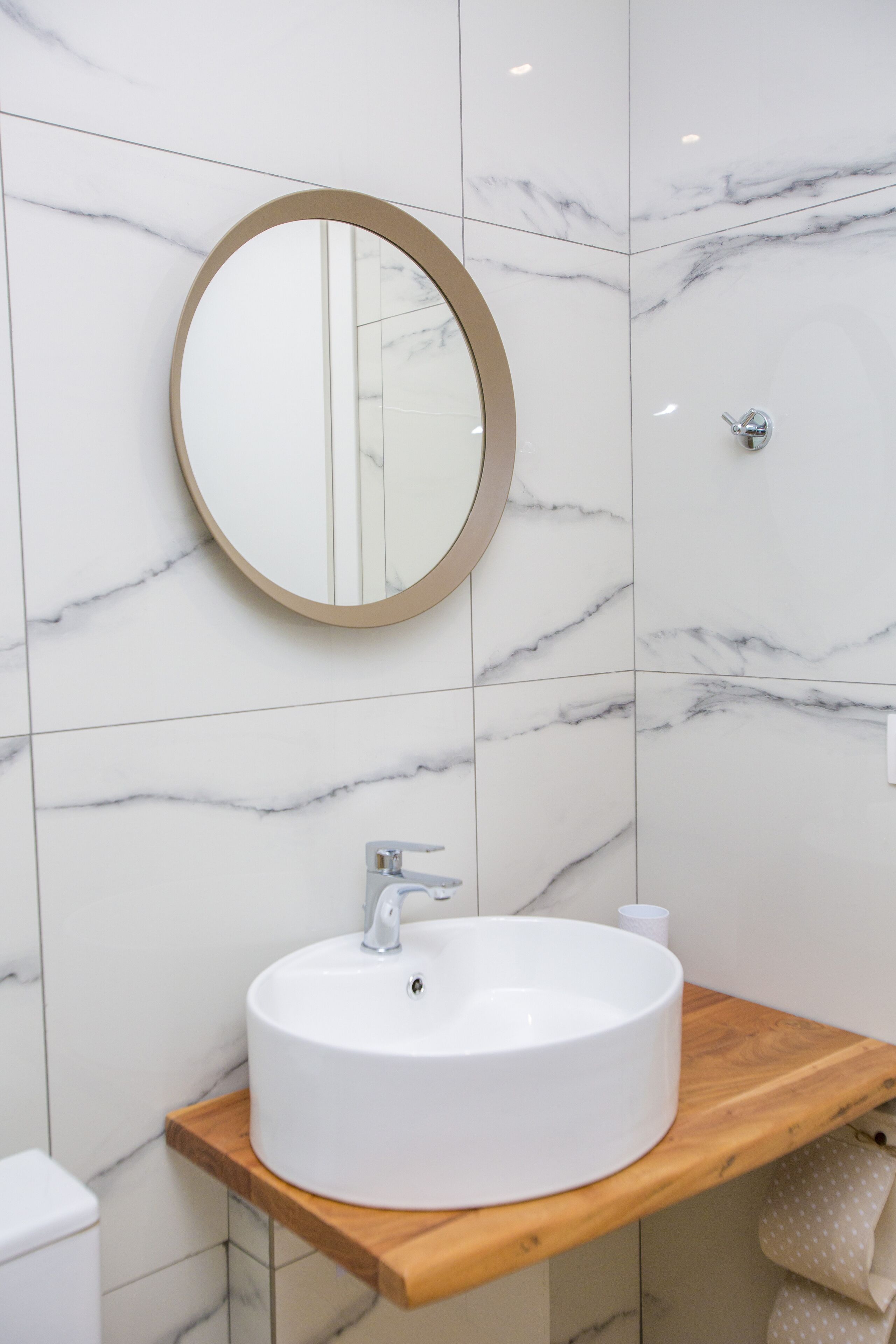 A bathroom with a marble wall, a round mirror, a white sink, and a wooden countertop.