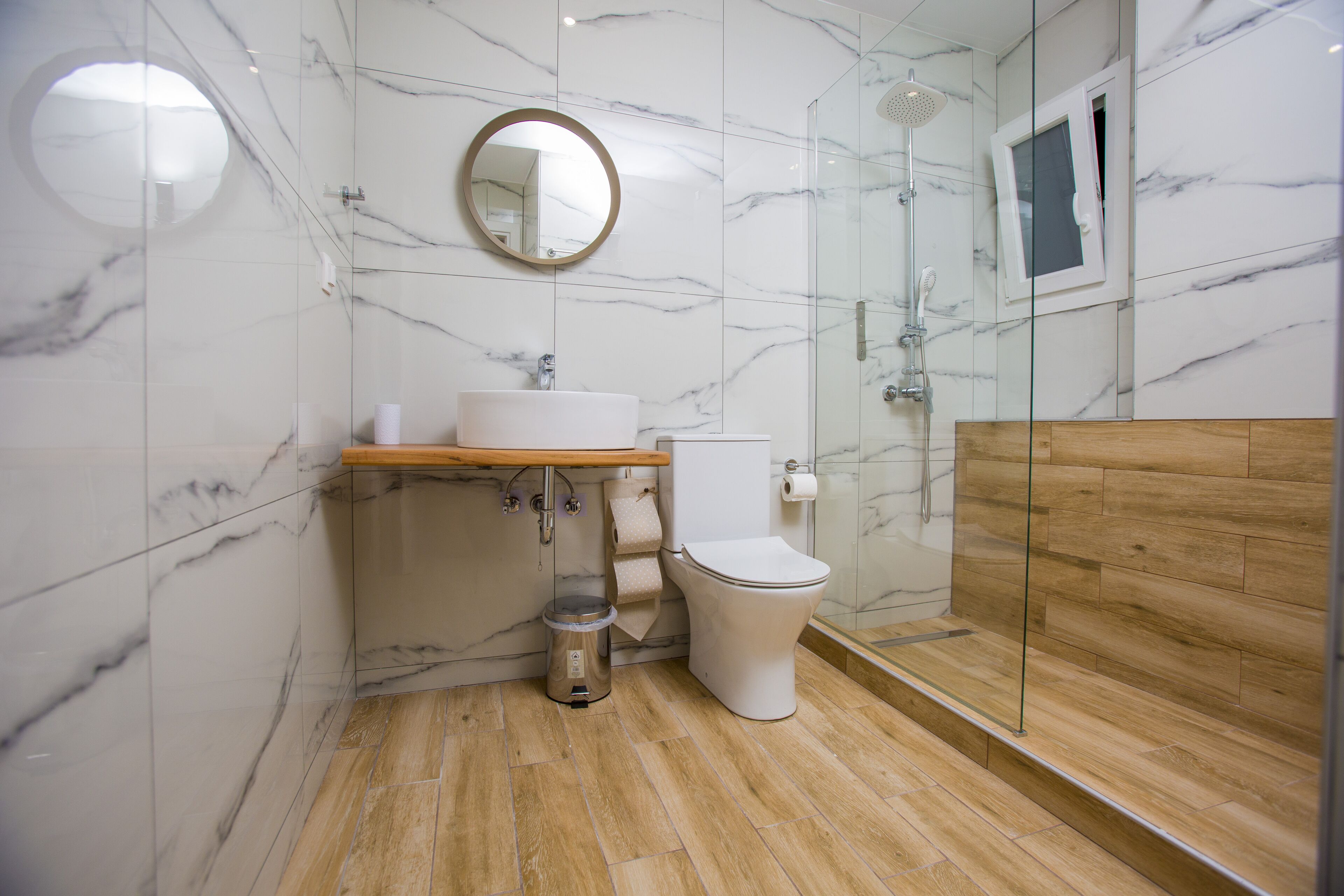 A modern bathroom with a marble wall, a wooden vanity, a toilet, and a glass shower enclosure.