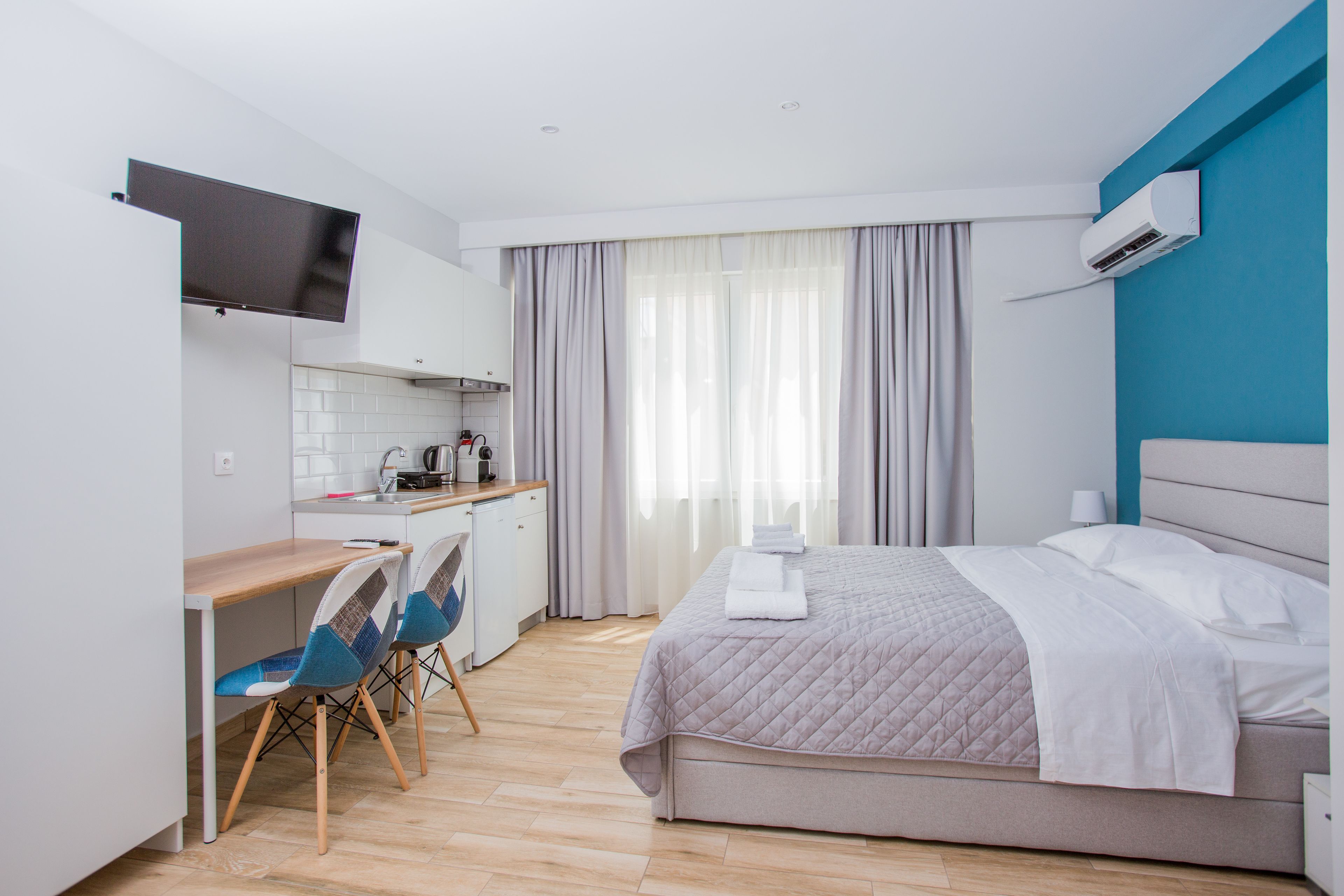 Moderni hotellihuone, jossa on sänky, työpöytä tuoleineen, pieni keittiö ja seinälle asennettu televisio.