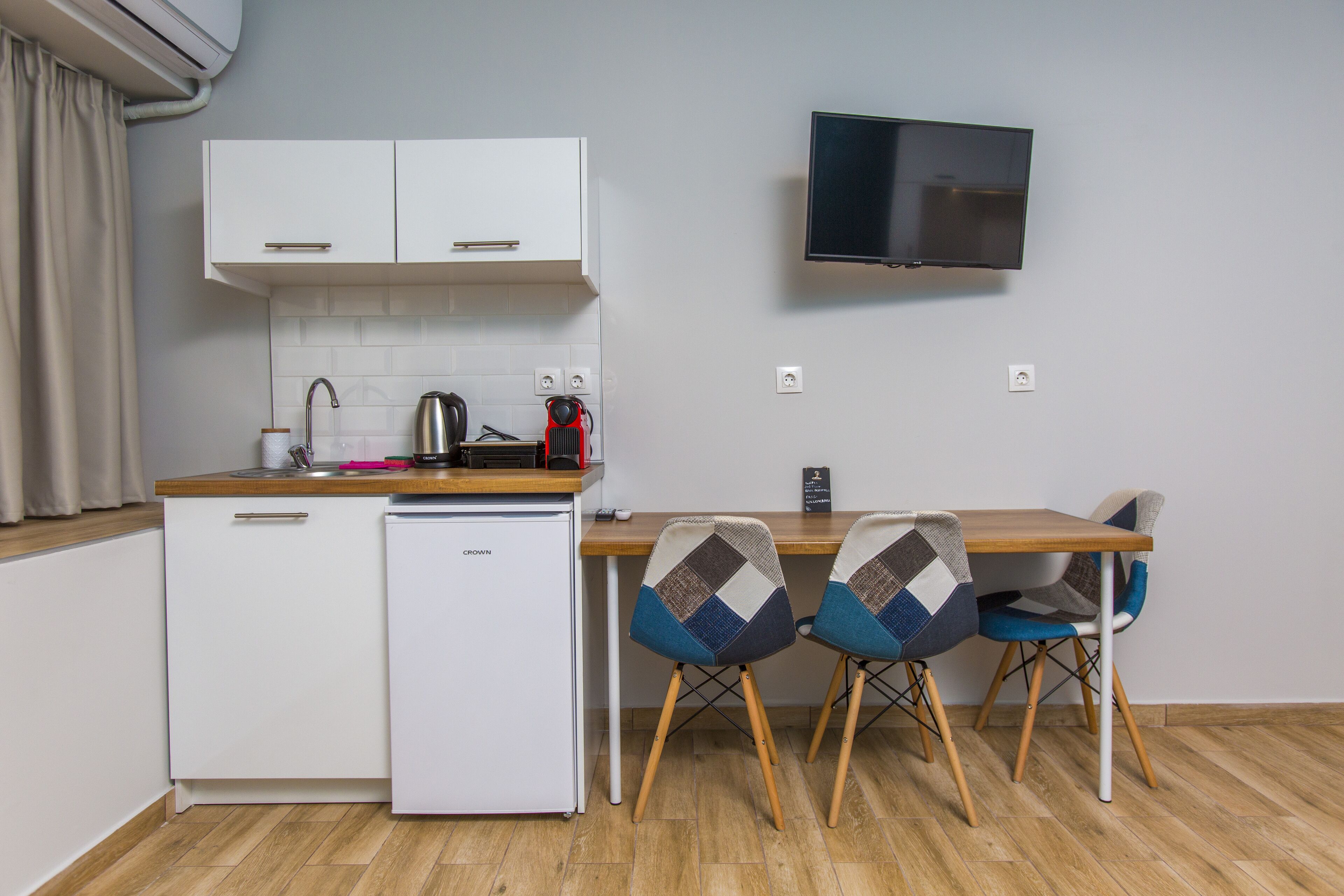 A compact kitchen with a refrigerator, a coffee maker, and a flat-screen TV mounted on the wall.