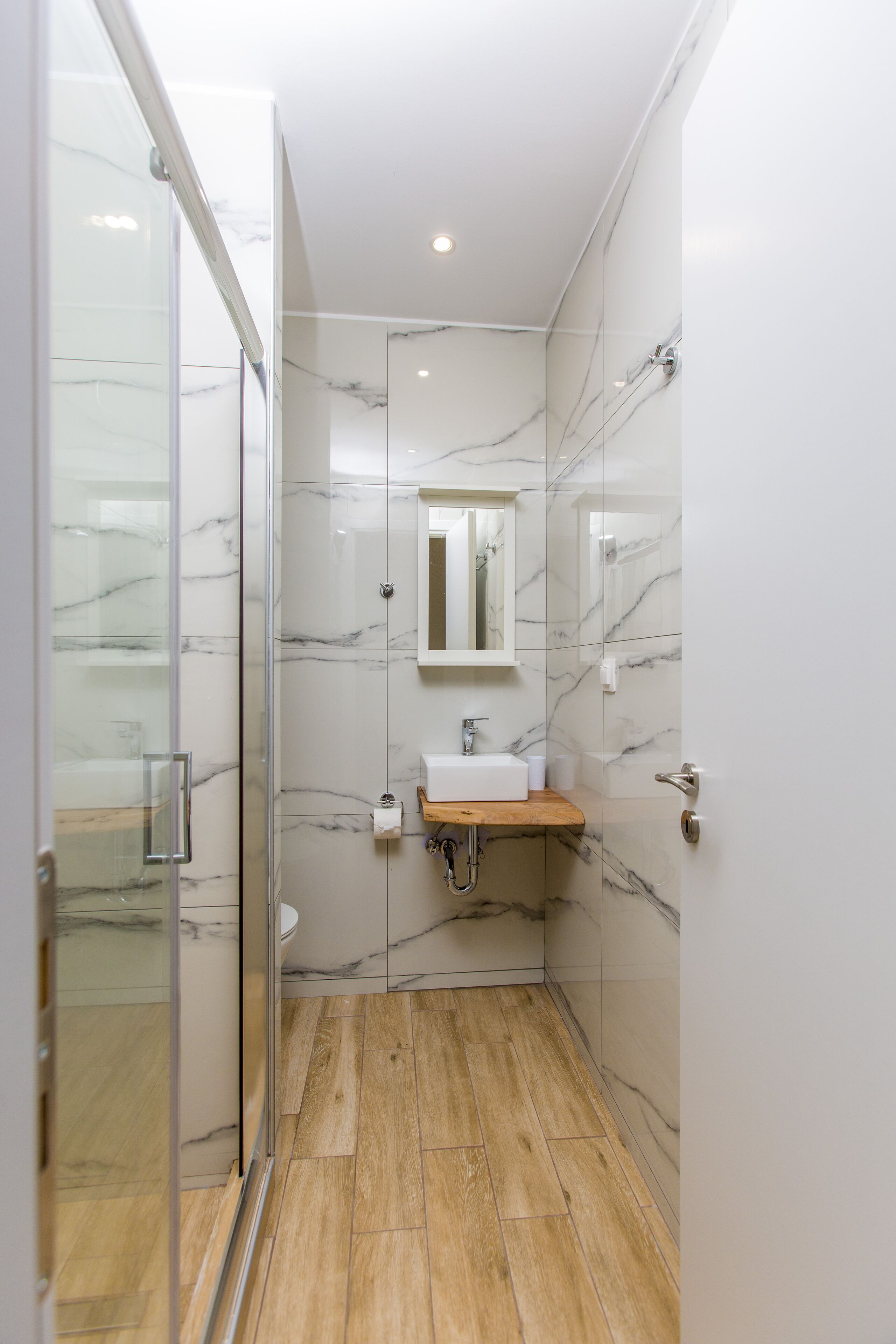 A modern bathroom with a glass shower enclosure, a wooden vanity, and marble walls.