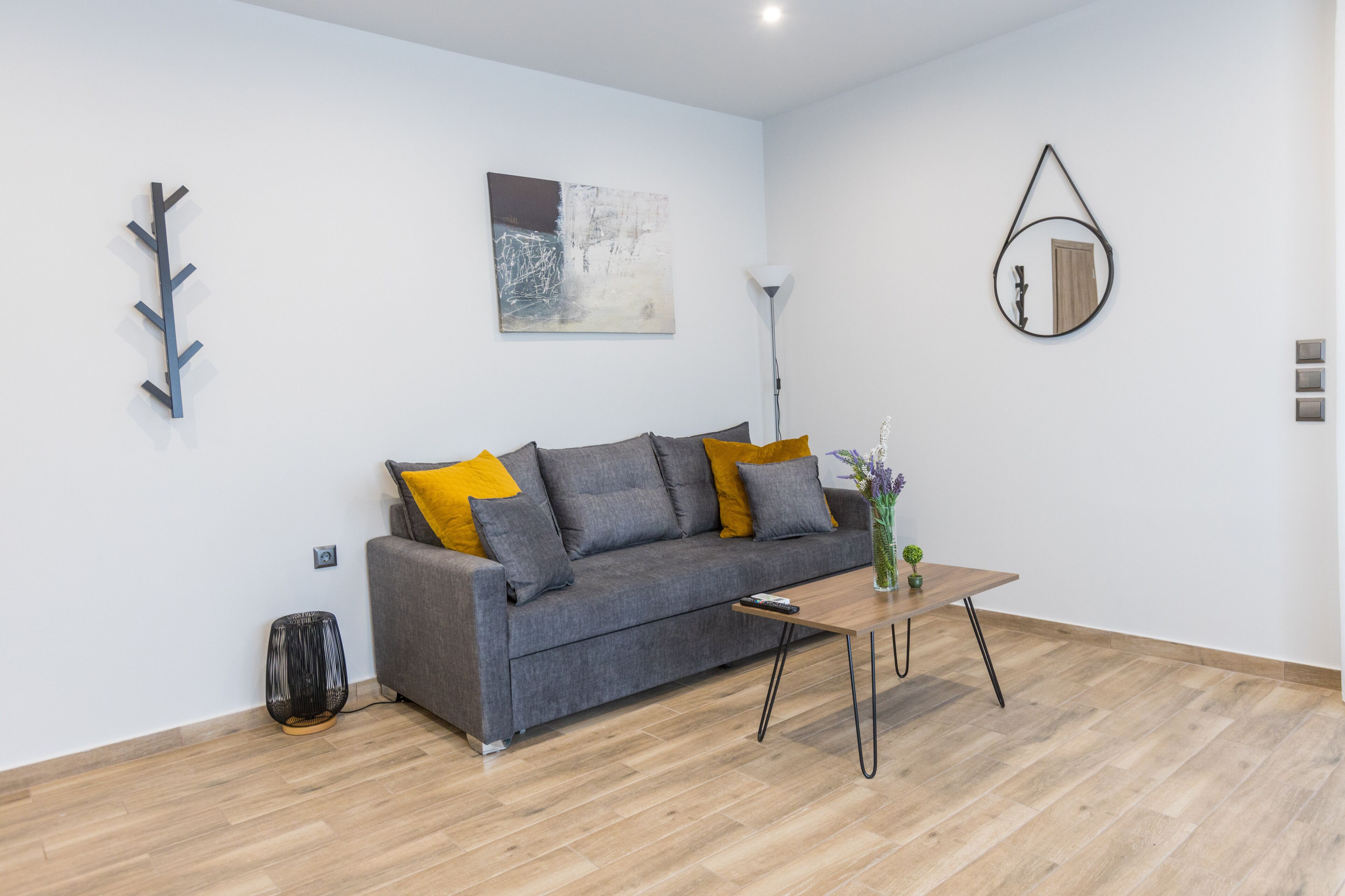 Una sala de estar moderna con un sofá gris, cojines amarillos, una mesa de centro de madera, una lámpara de pie, un espejo montado en la pared y una obra de arte abstracta.