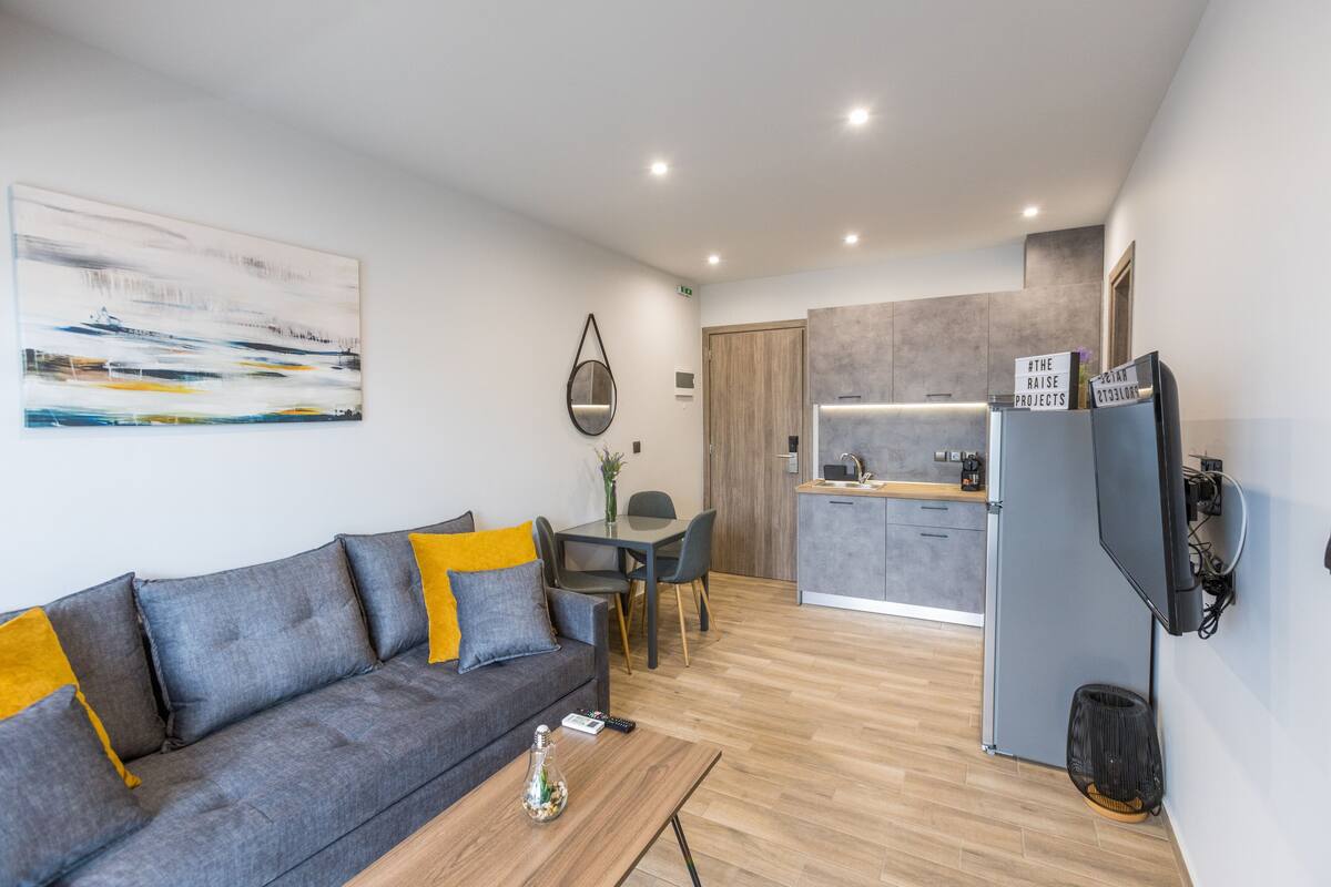 Una sala moderna con un sofá gris, una mesa de centro y un comedor con mesa y sillas. Se ve una cocina de fondo con gabinetes grises y un refrigerador.