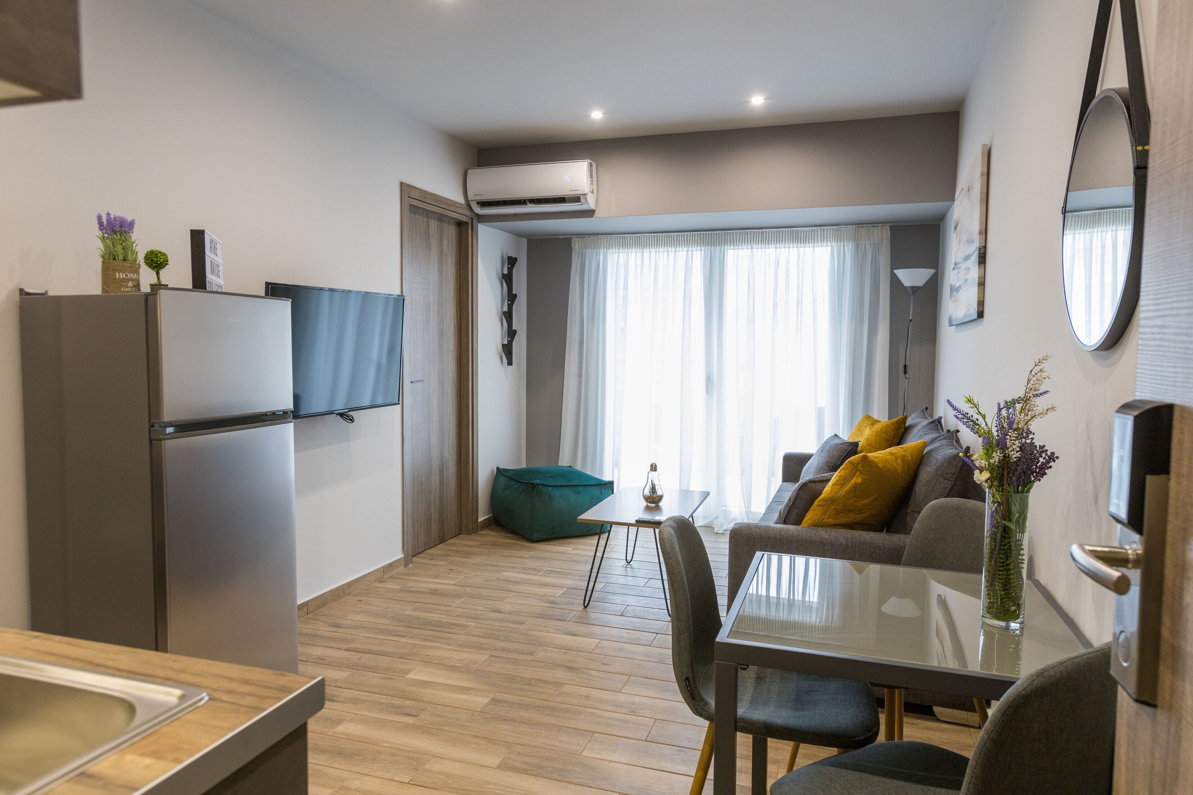 Un appartement moderne comprenant une cuisine, un coin repas et un salon. Il est équipé d’un téléviseur à écran plat, d’une table à manger avec des chaises, d’un canapé avec des coussins et d’une grande fenêtre avec des rideaux.