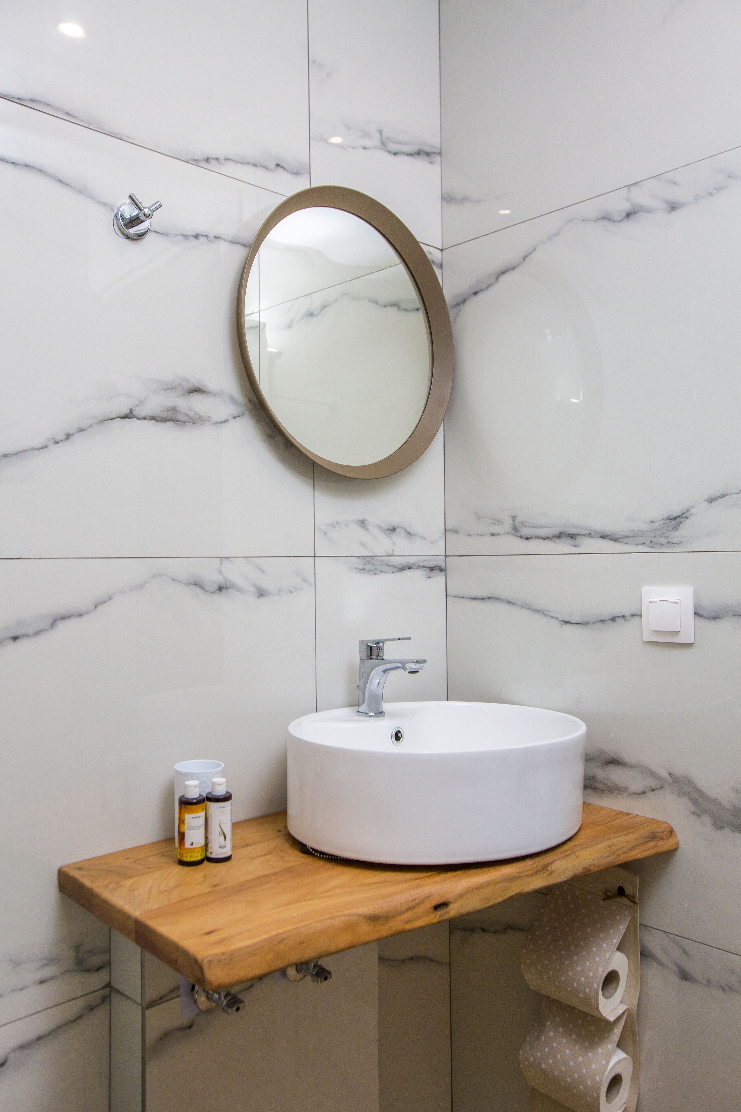 A bathroom with a marble wall, a round mirror, a white sink, and a wooden countertop.