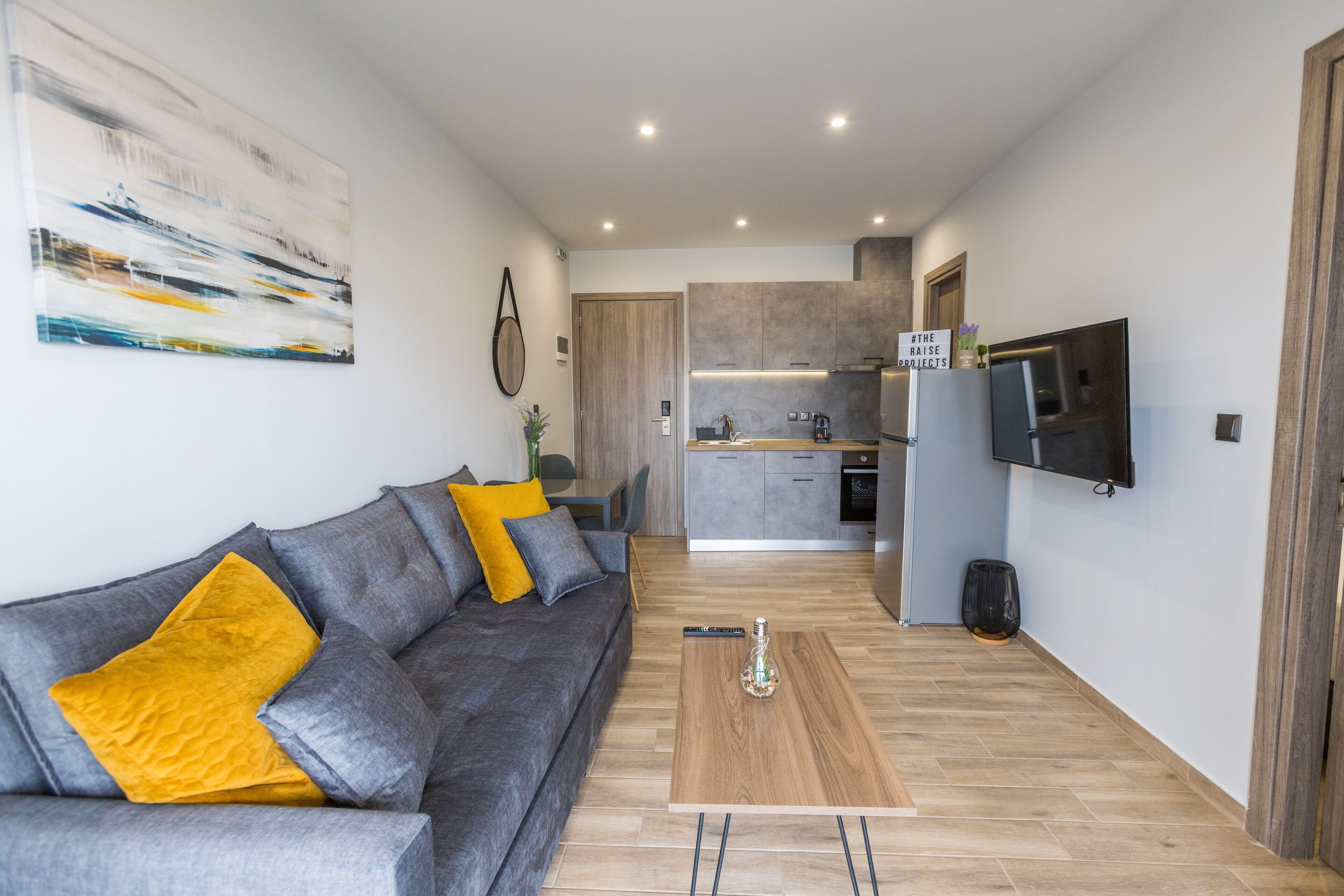 A modern living room with a grey sofa, a wooden coffee table, and a kitchen in the background.