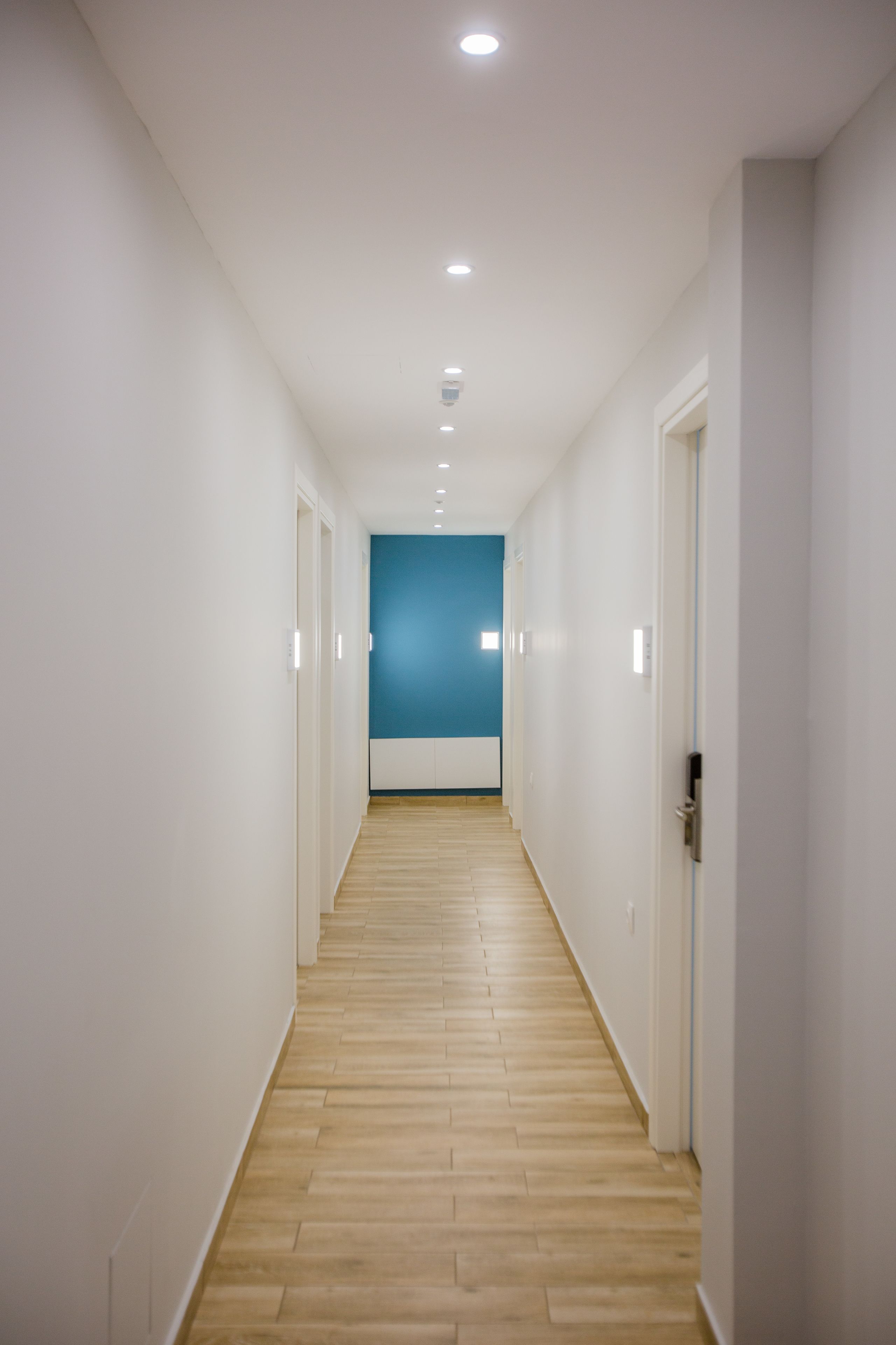 Un pasillo estrecho y bien iluminado, con suelo de madera y paredes blancas, que conduce a una habitación con un detalle en azul.