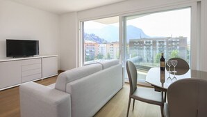 A modern living room with a sofa, a TV, a dining table with chairs, and a large window offering a view of buildings and mountains. - Roggia Apartments (Lugano)