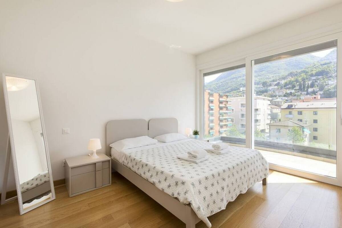 A bedroom with a bed, a bedside table, a mirror, and a large window with a view of buildings and hills.