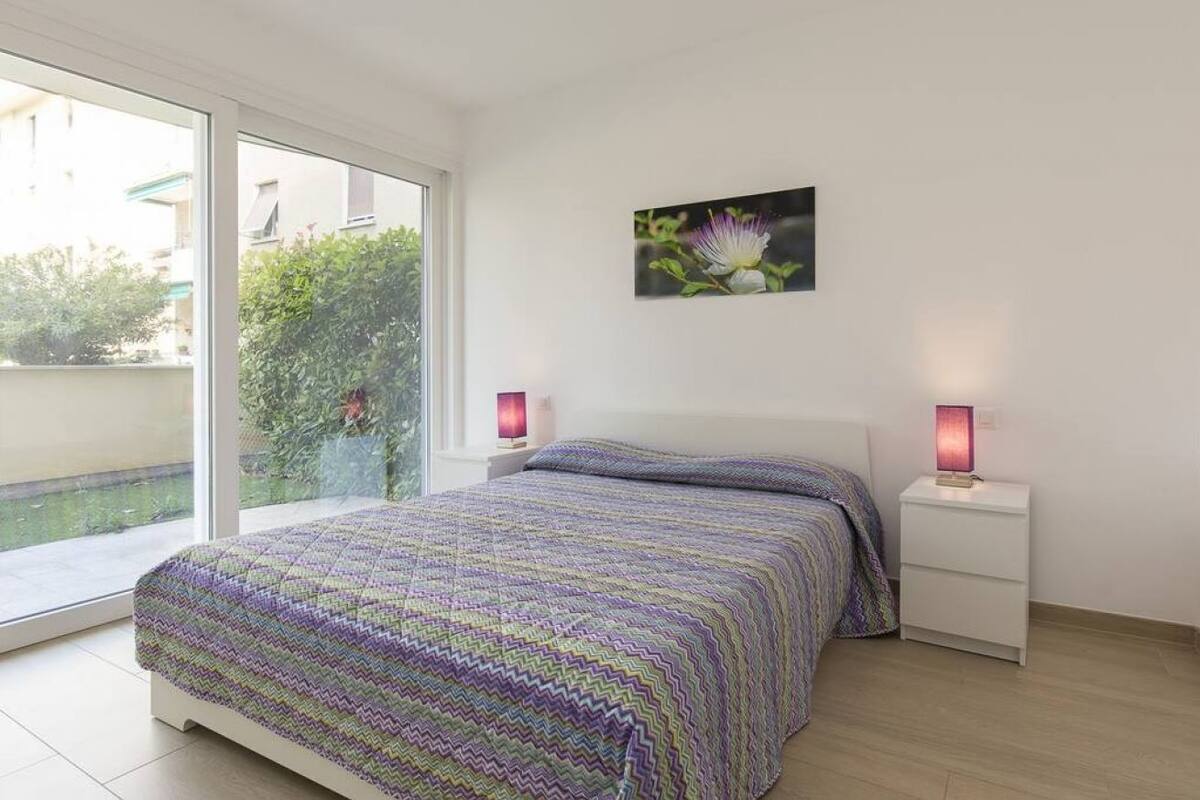 A bedroom with a large window, a bed with a patterned bedspread, a nightstand with a lamp, and a wall-mounted picture of a flower.